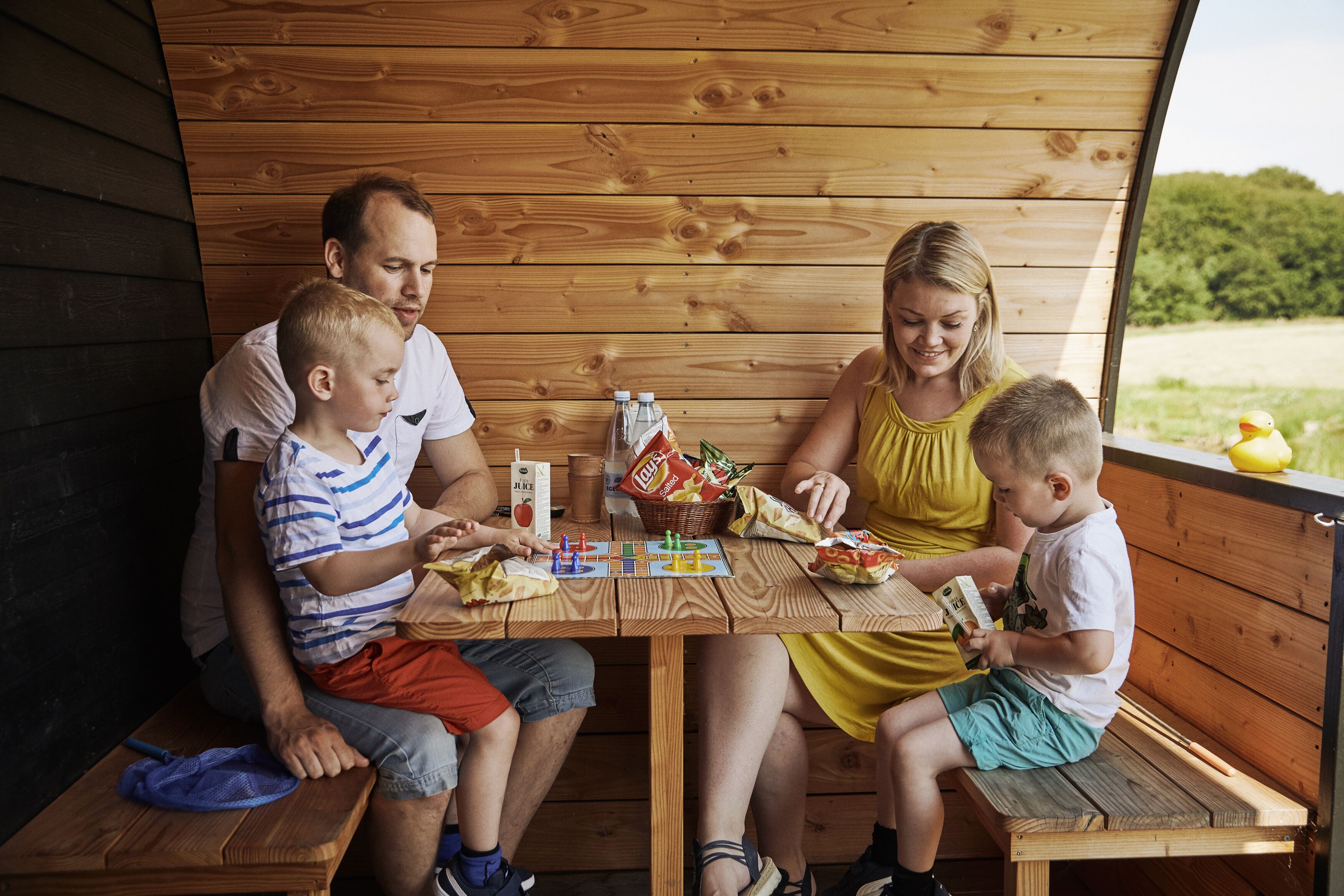A family of four seated at a wooden table with snacks and drinks, in a wooden cabin-like setting.