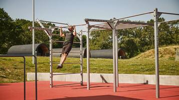 A man performing pull-ups on a metal bar structure in an outdoor setting with a red floor and greenery in the background.