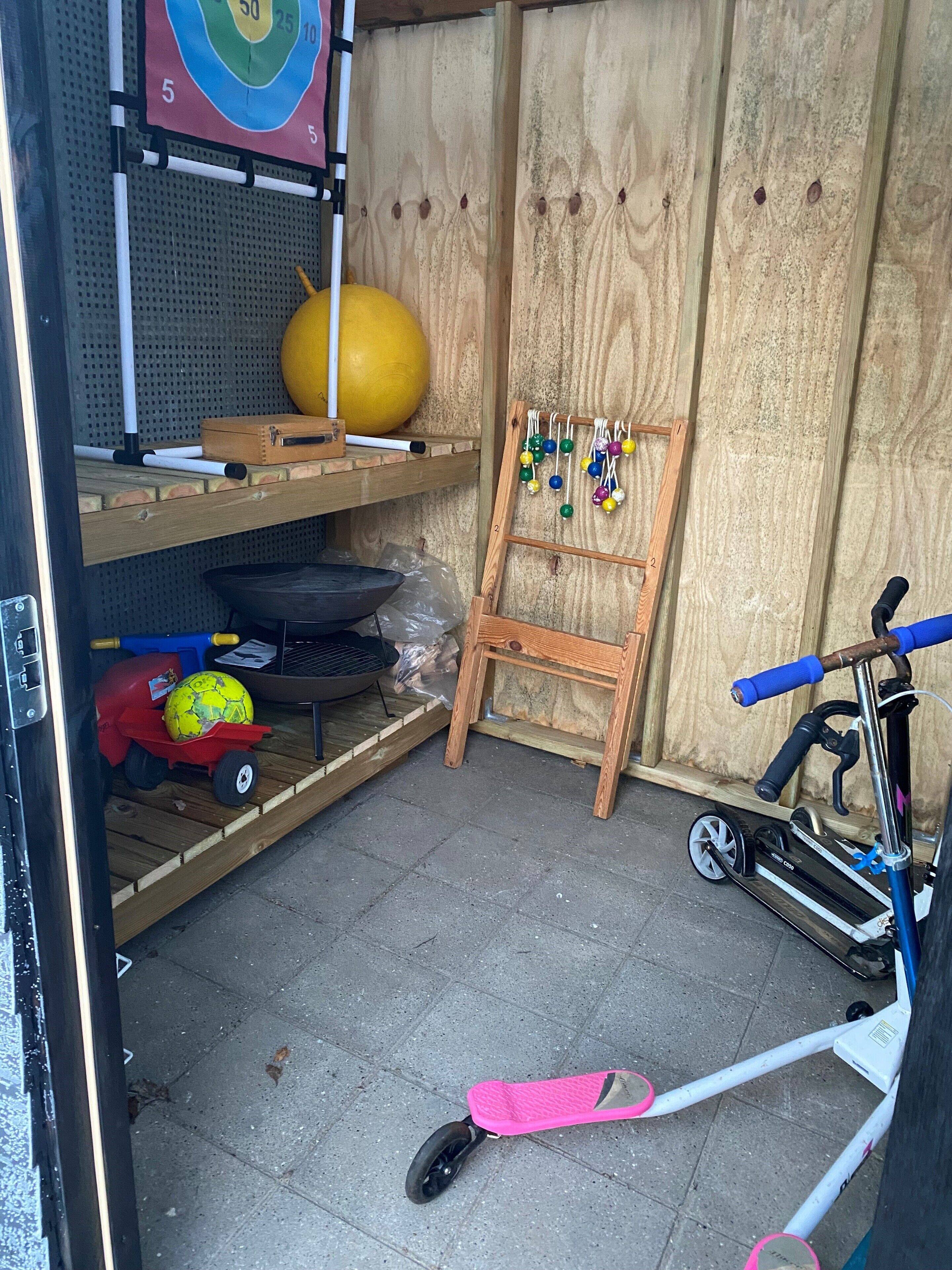 A room with a wooden floor, a target on the wall, a yellow ball, a small table with a ball, a scooter, and a pink skateboard.
