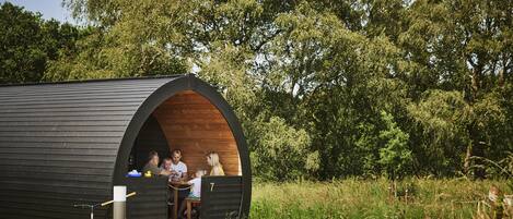 Une structure en bois surmontée d’une arche semi-circulaire, deux personnes assises à l’intérieur, et une table sur laquelle se trouvent divers objets.