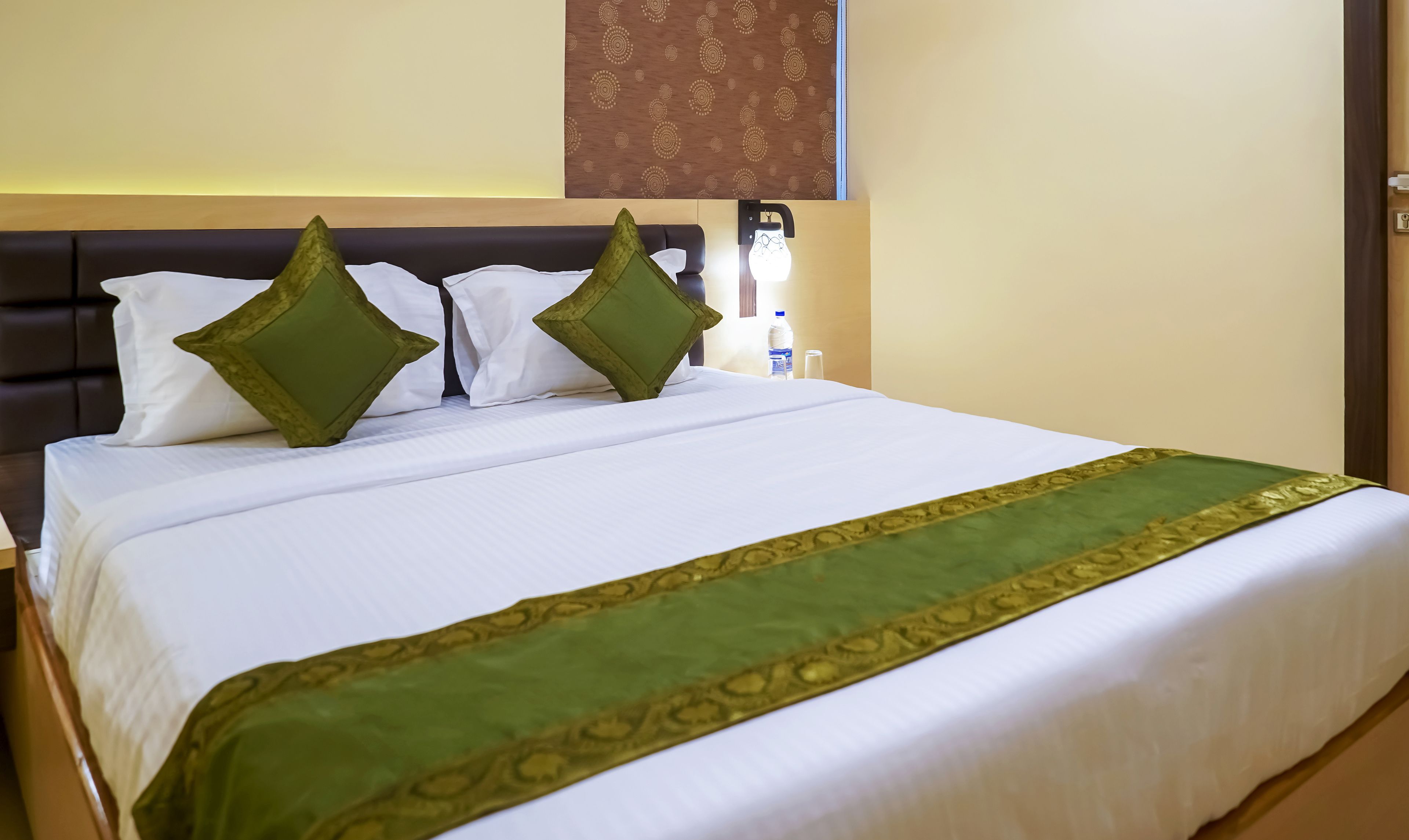 A neatly made bed with white linens and a green decorative border, two green pillows, a bottle of water, and a wall-mounted light fixture.