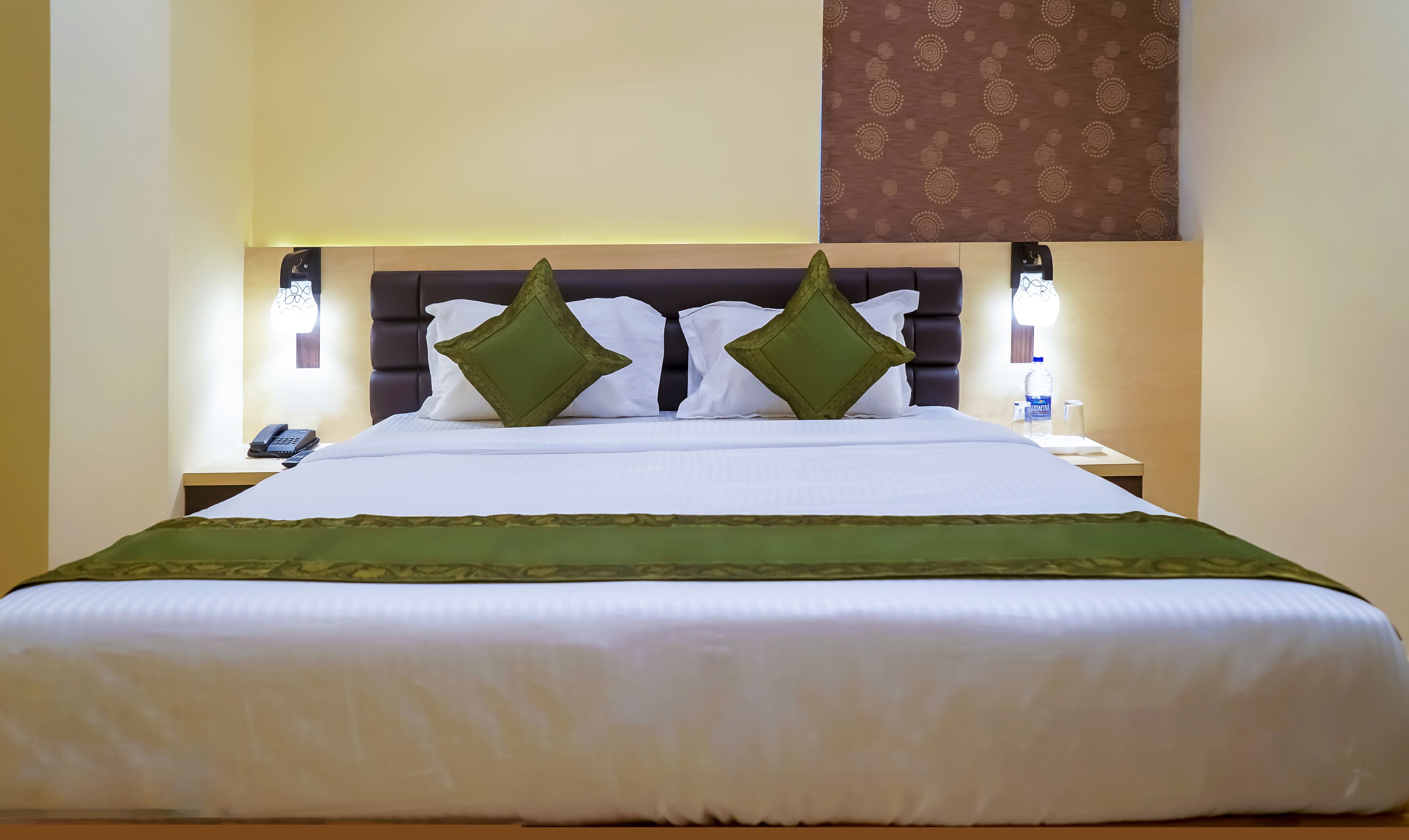 A neatly made bed with a green bedspread and two green pillows, a bedside table with a phone and a bottle of water, and a wall-mounted light fixture.