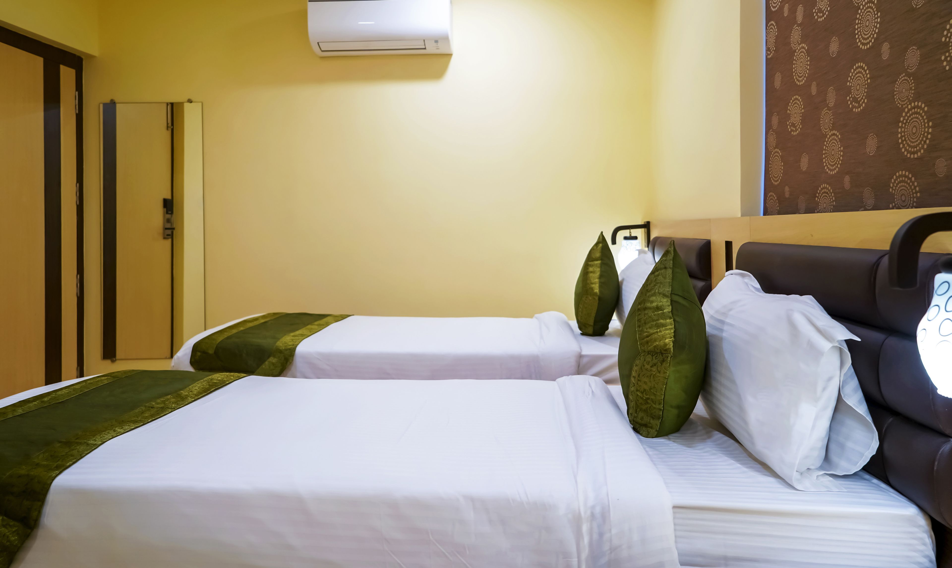 A hotel room with two beds, each with white linens and green pillows. There is a wall-mounted air conditioner above the beds, and a patterned curtain on the window.