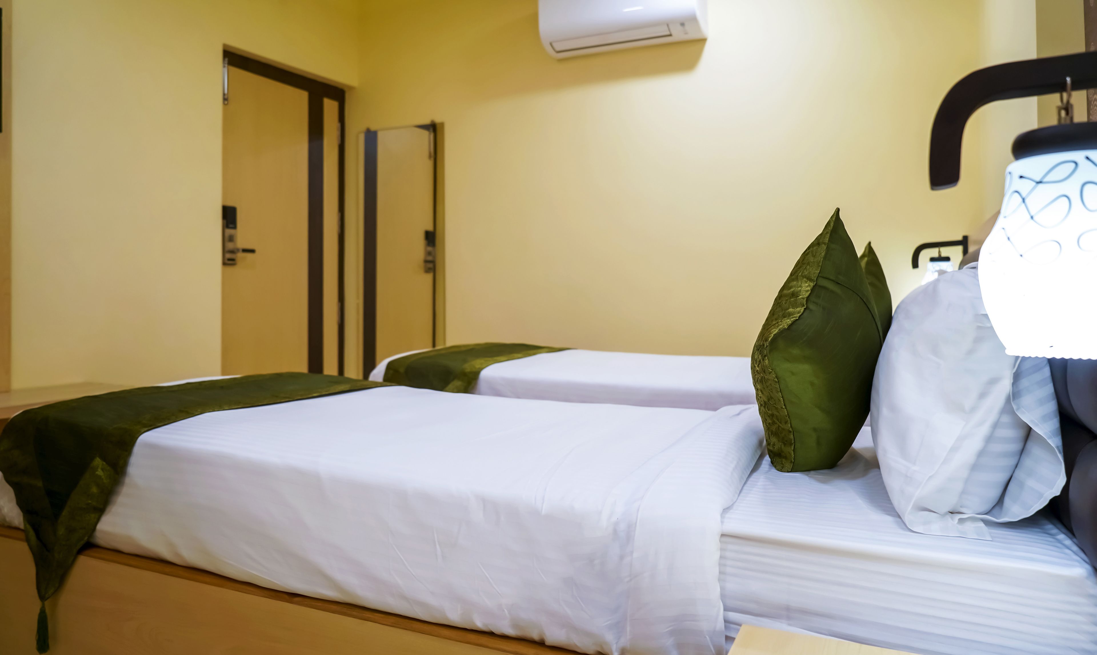 A hotel room with two beds, each with white linens and green pillows. There is a door and an air conditioning unit on the wall.
