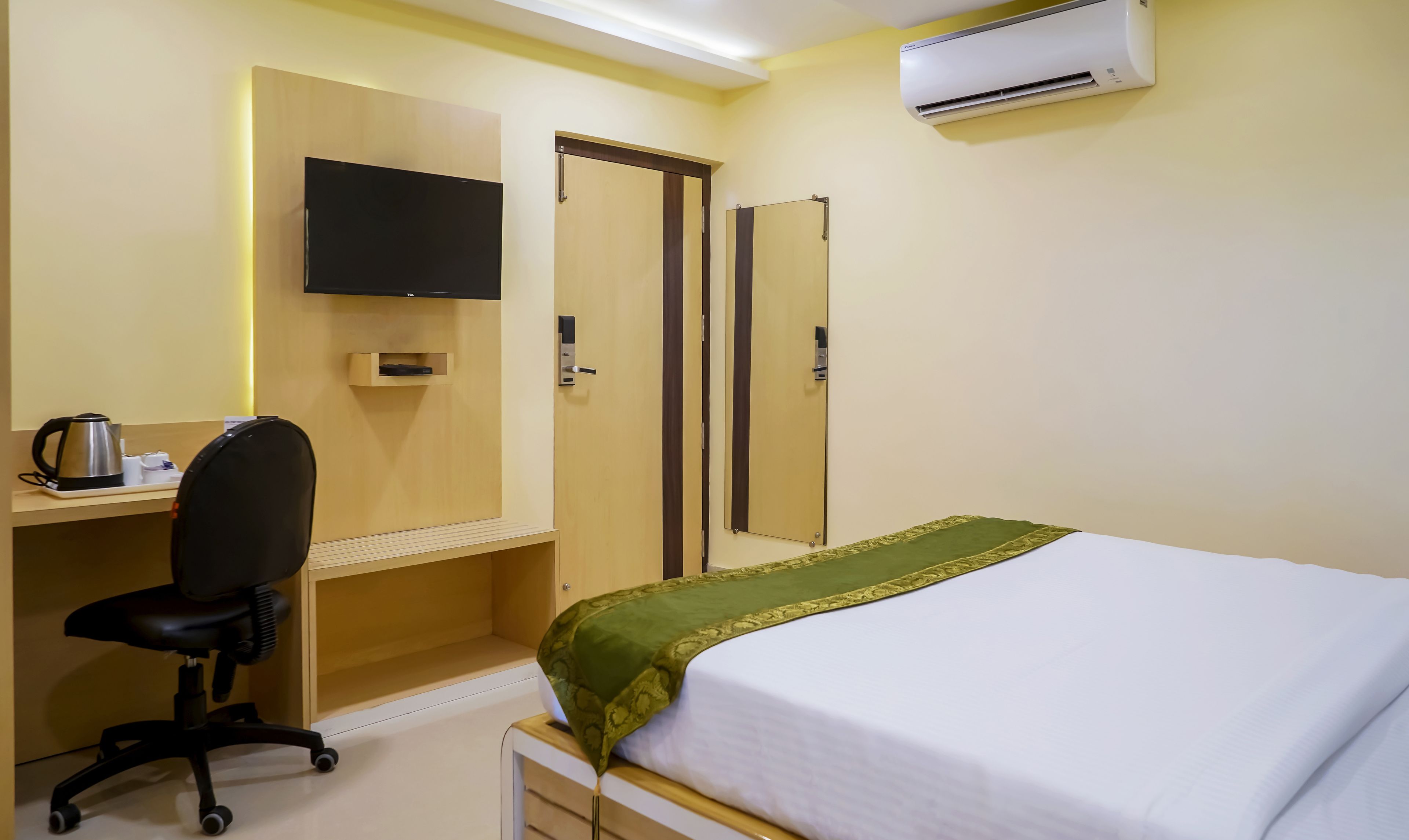 A hotel room with a bed, a desk with a chair, a television, a wardrobe, and an air conditioning unit.