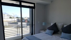 A bedroom with a large bed, a nightstand, a lamp, and a view of a cityscape through a sliding door. - Two Bedroom Apartment (Cape Town)
