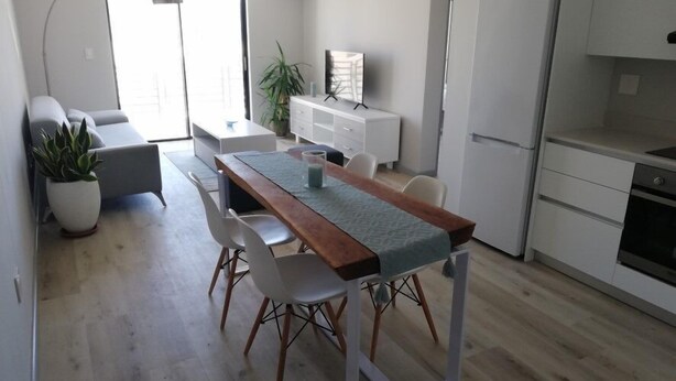 A modern kitchen with a wooden dining table, white chairs, and a refrigerator. - Two Bedroom Apartment (Cape Town)