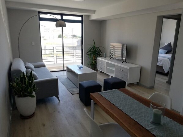 A modern living room with a sofa, coffee table, and a dining area with a bench. There is a large window with a view of the outside.