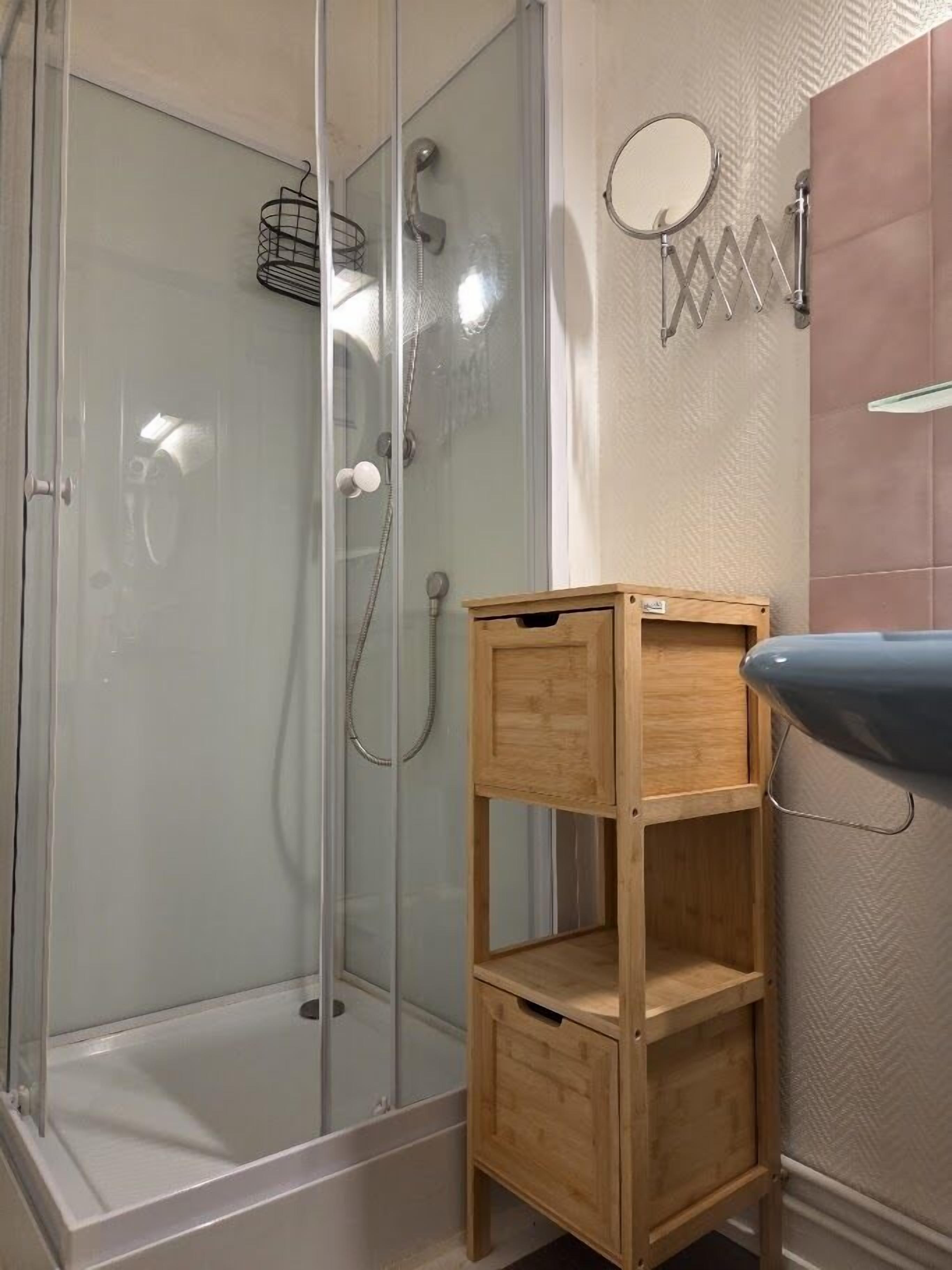 A bathroom with a glass-enclosed shower, a wooden shelf unit, a round mirror, and a sink.