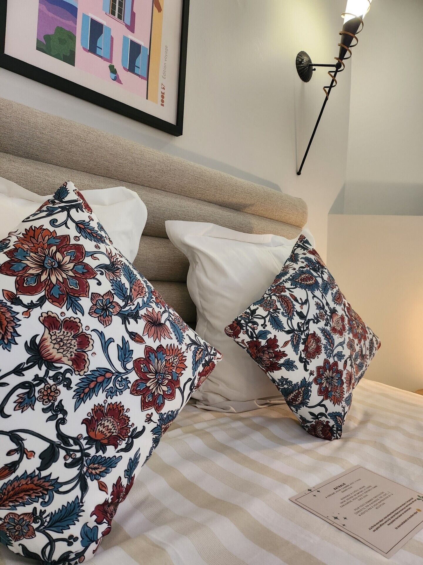 Una cama con ropa de cama a rayas, dos almohadas estampadas con motivos florales, una obra de arte enmarcada sobre la cama y una lámpara fijada a la pared.