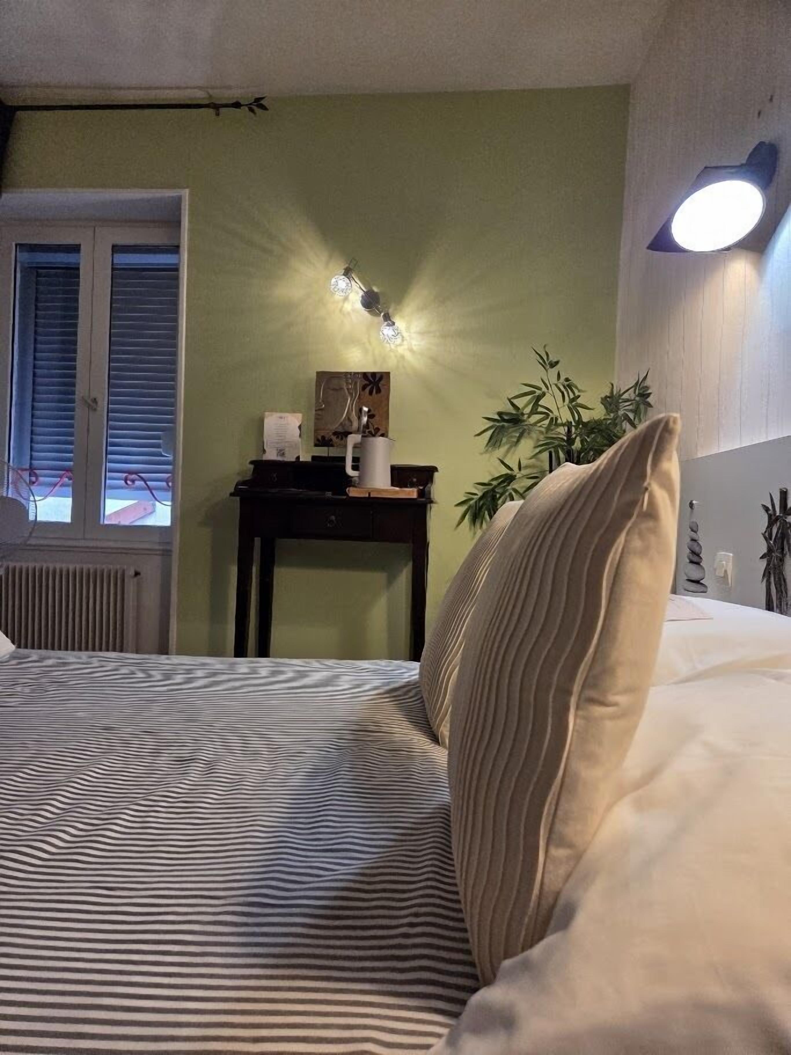 Et soveværelse med seng, natbord, vægmonteret lampe, et vindue med rullegardiner og en radiator.
