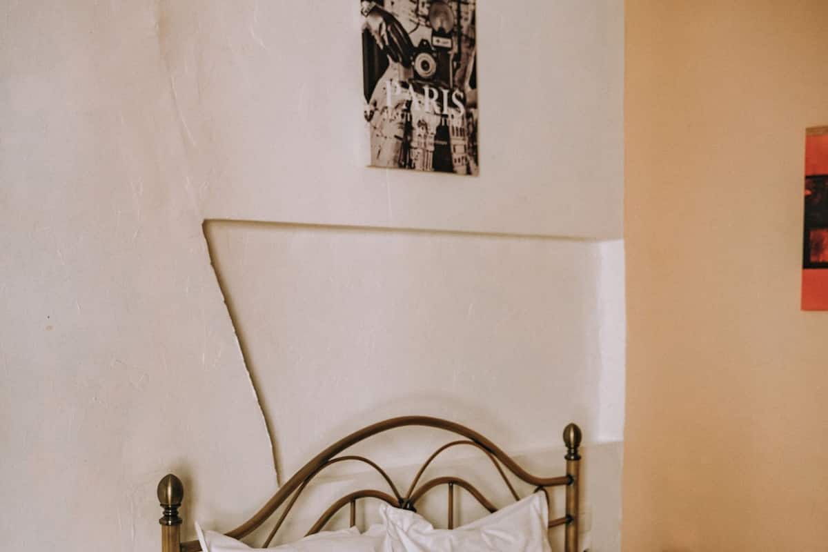 Una cama con cabecera, un taburete de madera redondo, una lámpara de pie y un cartel en la pared.
