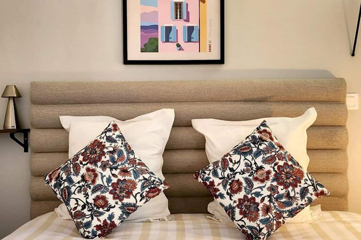 Una cama con dos almohadas, cabecera, lámpara y un cuadro en la pared.