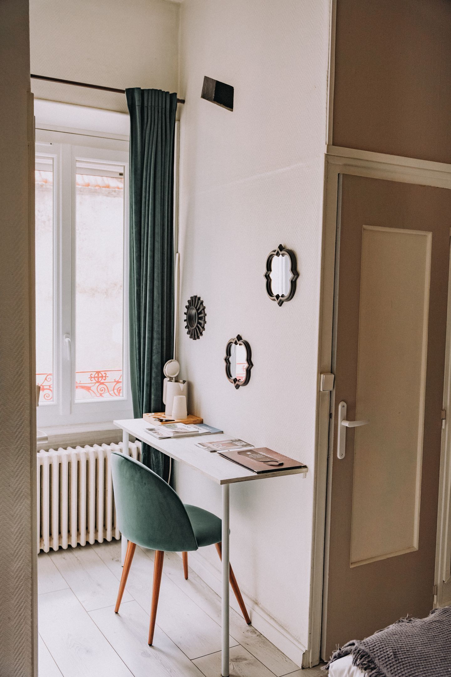Ein Zimmer mit weißem Schreibtisch, grünem Stuhl, Heizkörper, einem Fenster mit Vorhängen und einer Tür.