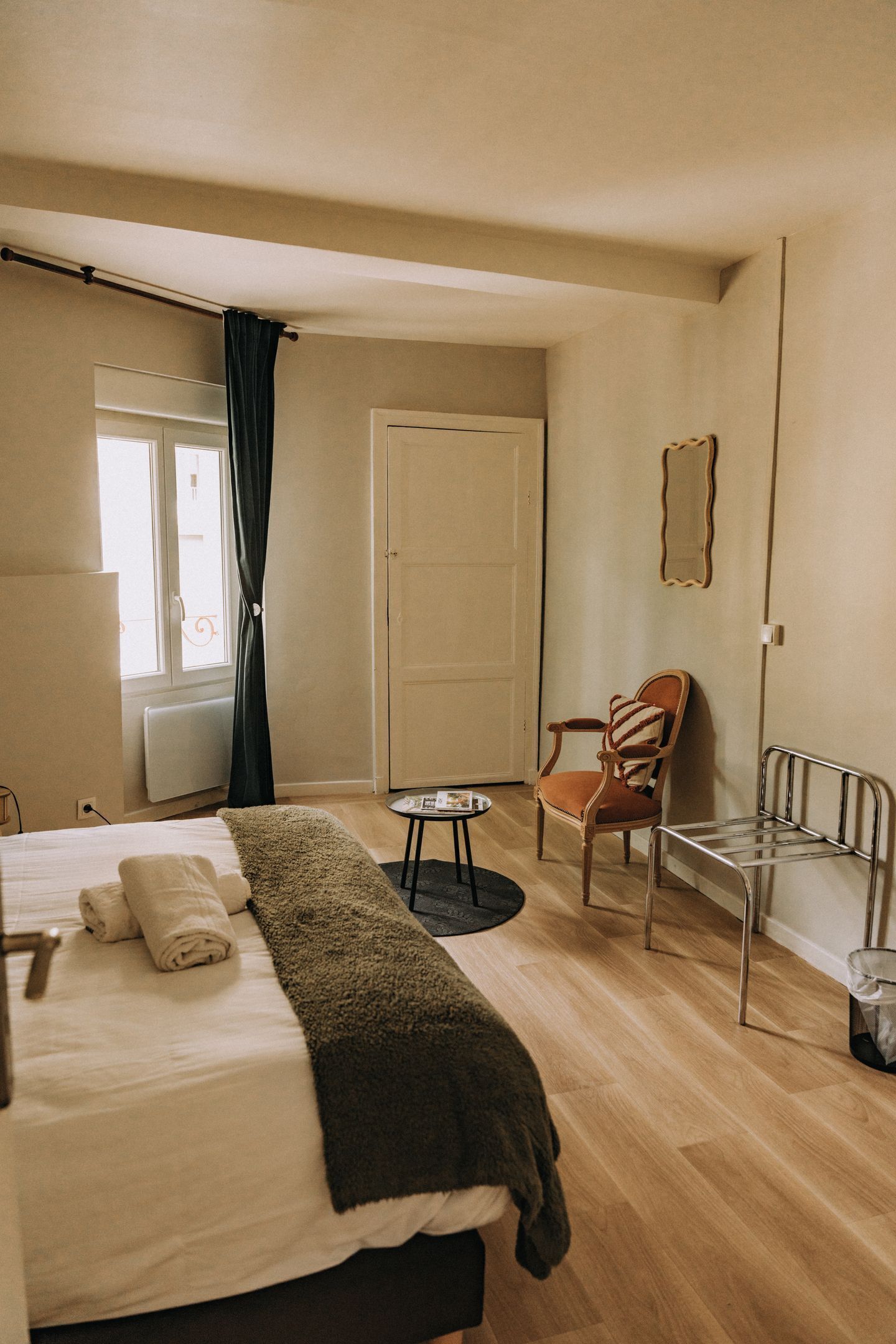 A hotel room with a bed, a chair, a small table, a radiator, a window with curtains, and a door.