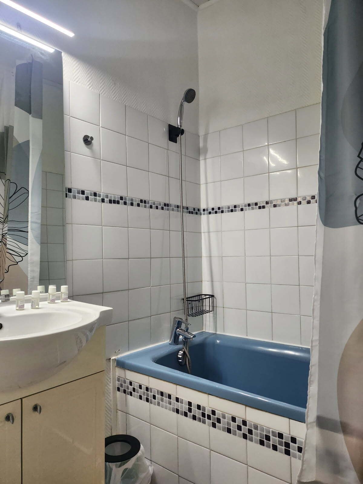 A bathroom with white tiled walls, a blue bathtub, a white sink with toiletries, and a shower head.