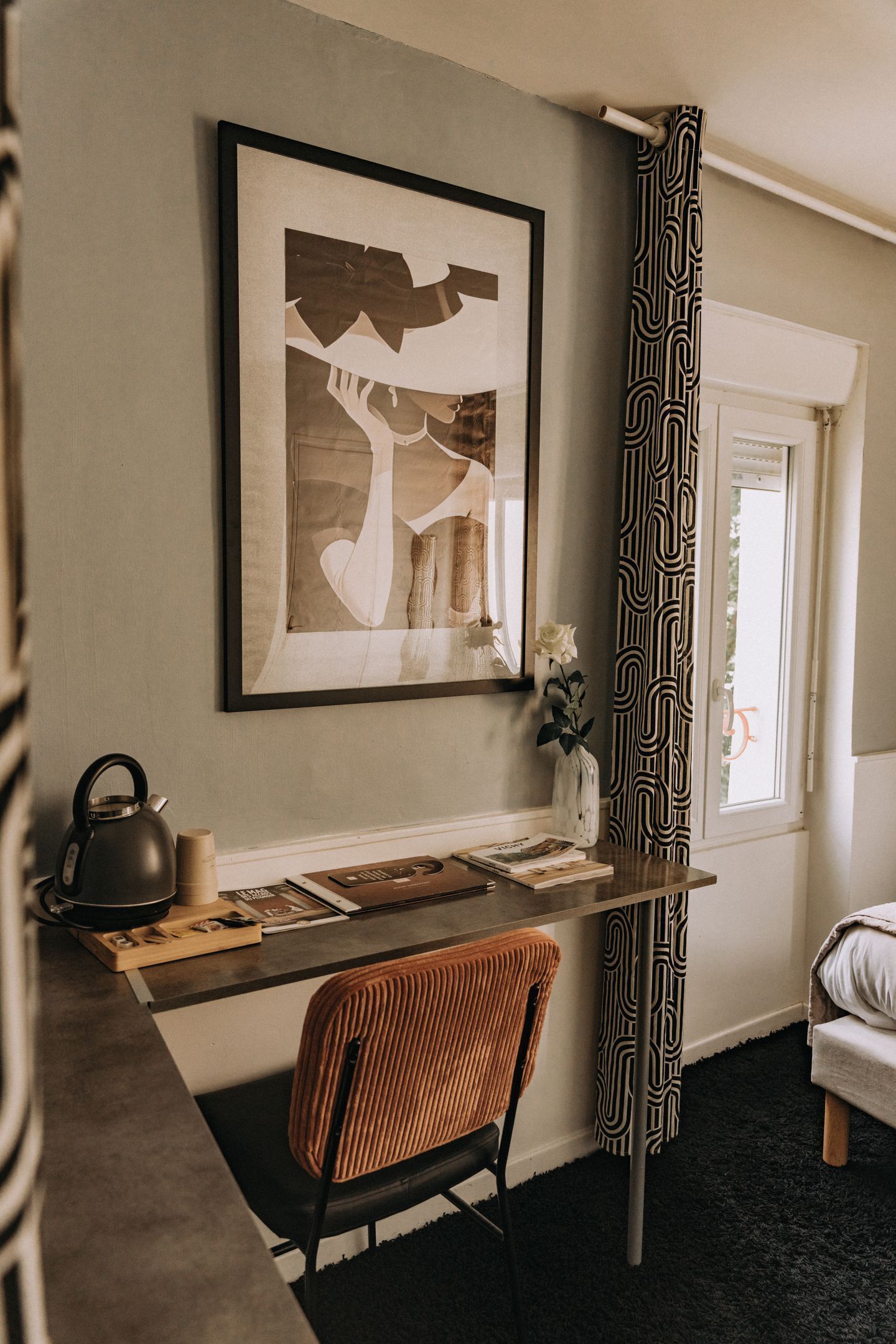 Una habitación con una obra de arte enmarcada, un escritorio con un hervidor, una silla y una ventana con cortinas estampadas.