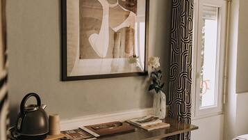 Una habitación con una obra de arte enmarcada, un escritorio con un hervidor, una silla y una ventana con cortinas estampadas.