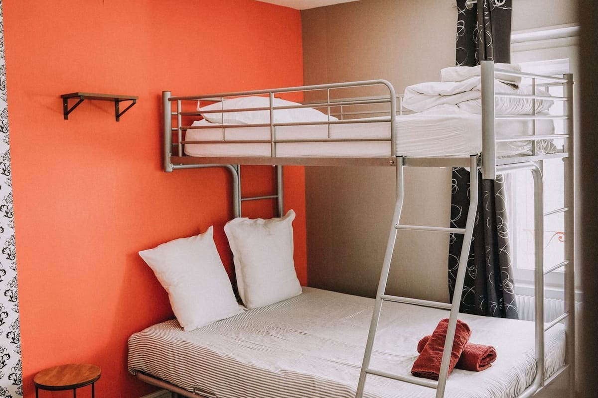 Dormitorio con literas, una pequeña mesa y una silla, paredes color naranja y una ventana con cortinas.