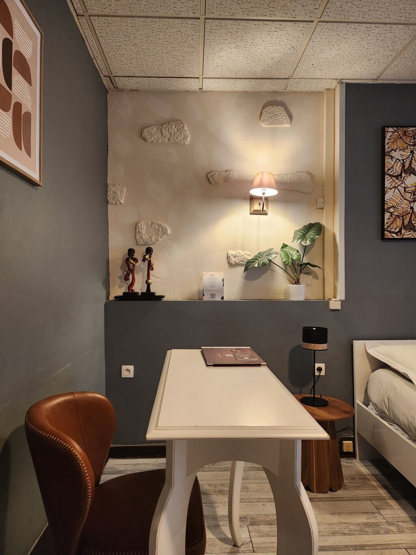 Una habitación compacta con una mesa blanca, un sillón de cuero marrón, una mesita de madera pequeña, una cama y una lámpara de pared.