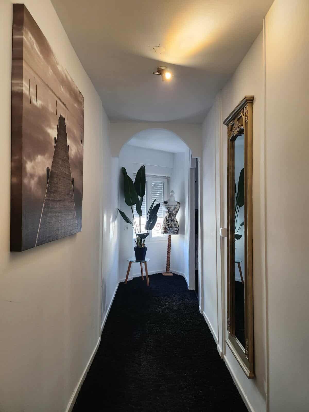 Un pasillo con alfombra oscura, un cuadro grande a la izquierda, una planta sobre una mesita y un espejo a la derecha.