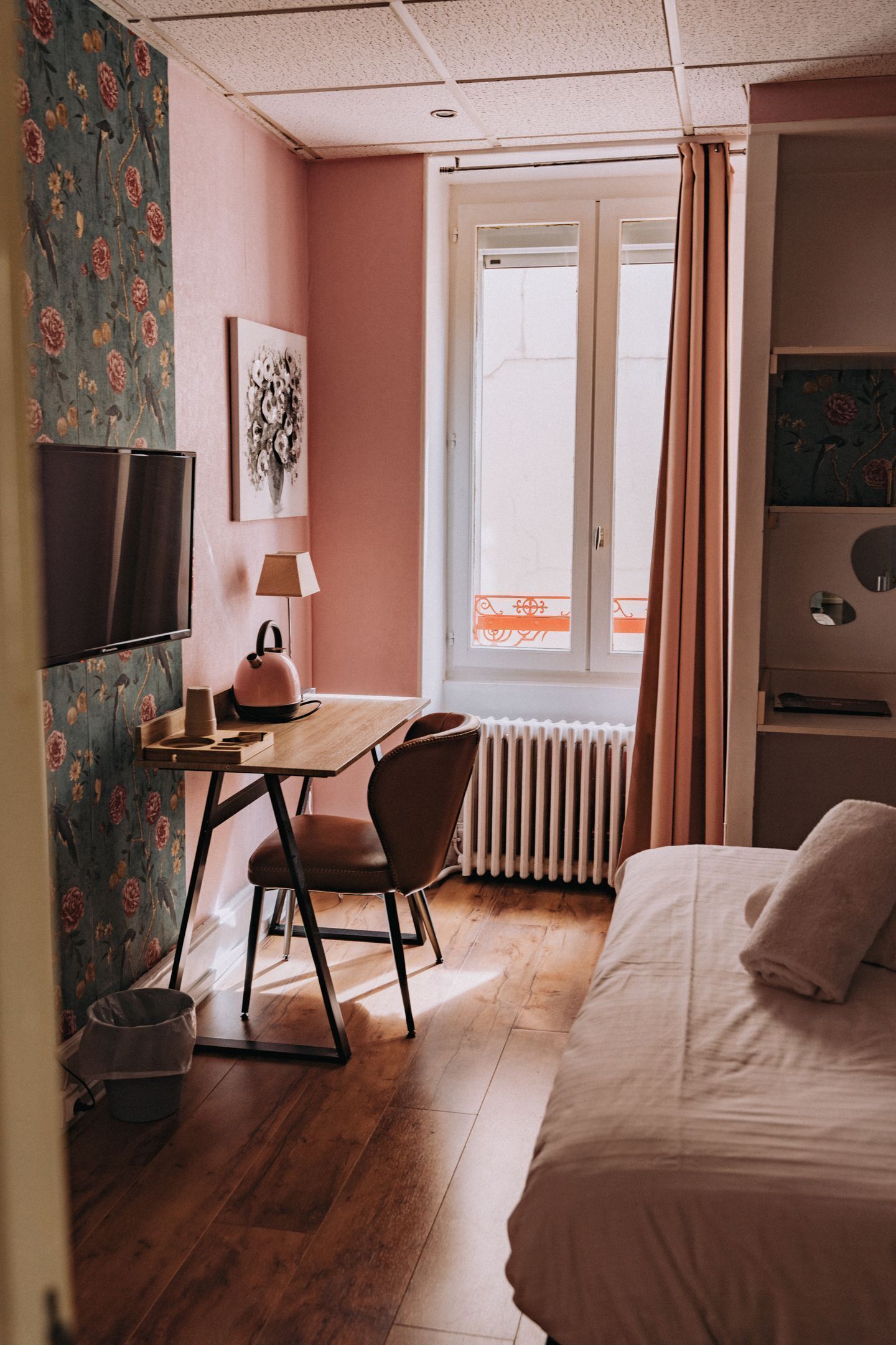A room with a bed, a desk with a chair, a radiator, a window with curtains, and a television on the wall.