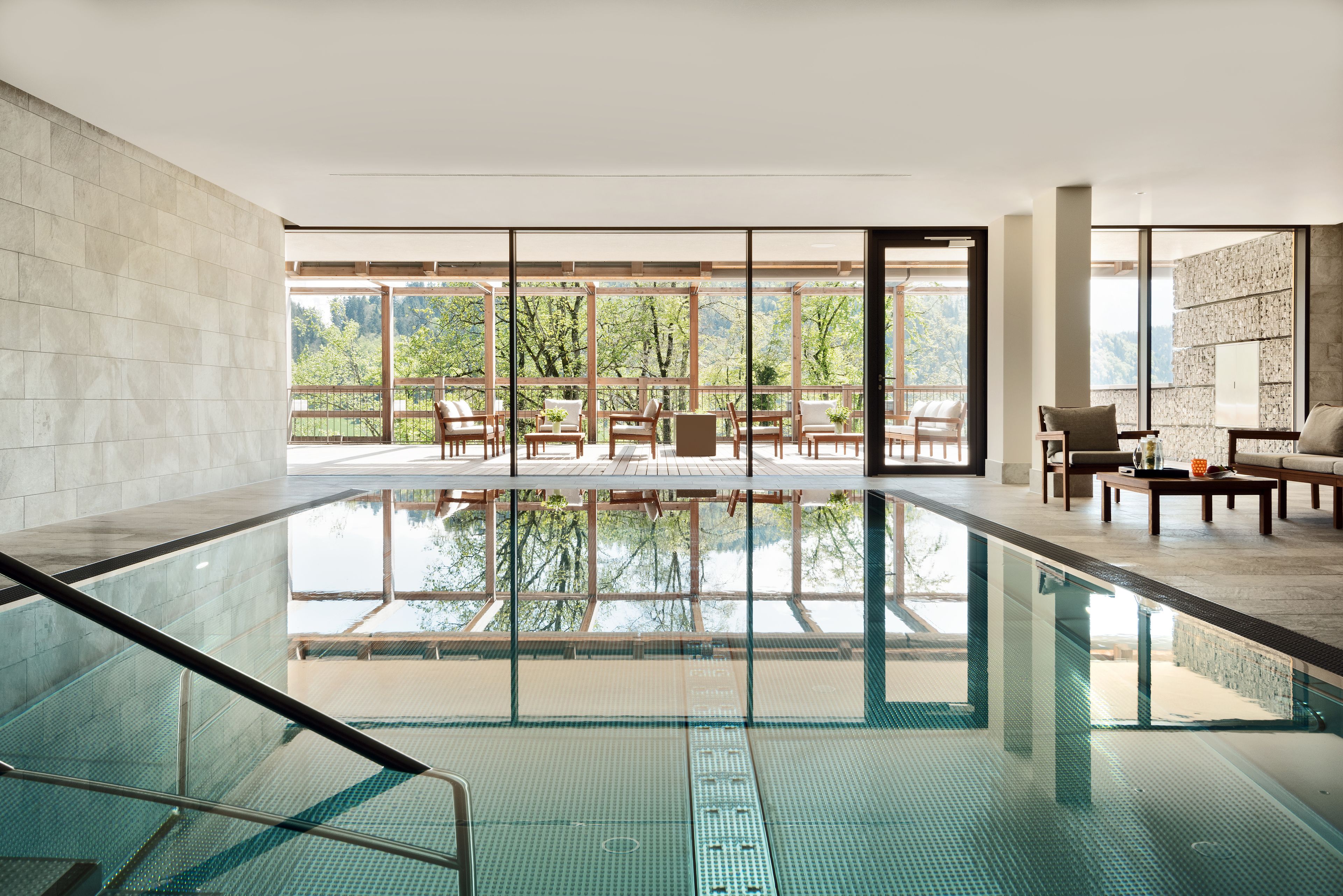 A modern indoor swimming pool with clear glass walls, a wooden deck area, and a seating area with tables and chairs.