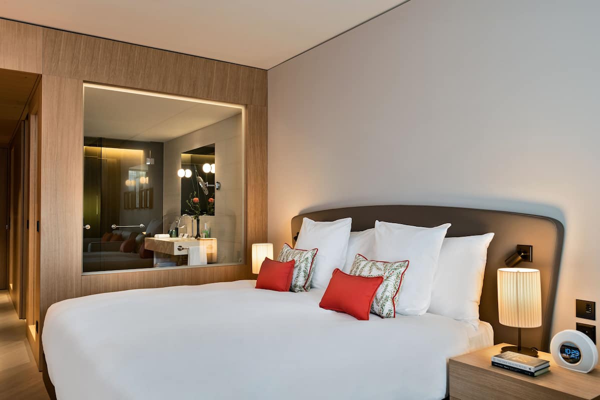 A modern hotel room with a large bed, a bedside table with a lamp, a digital clock, and a mirror reflecting the room.
