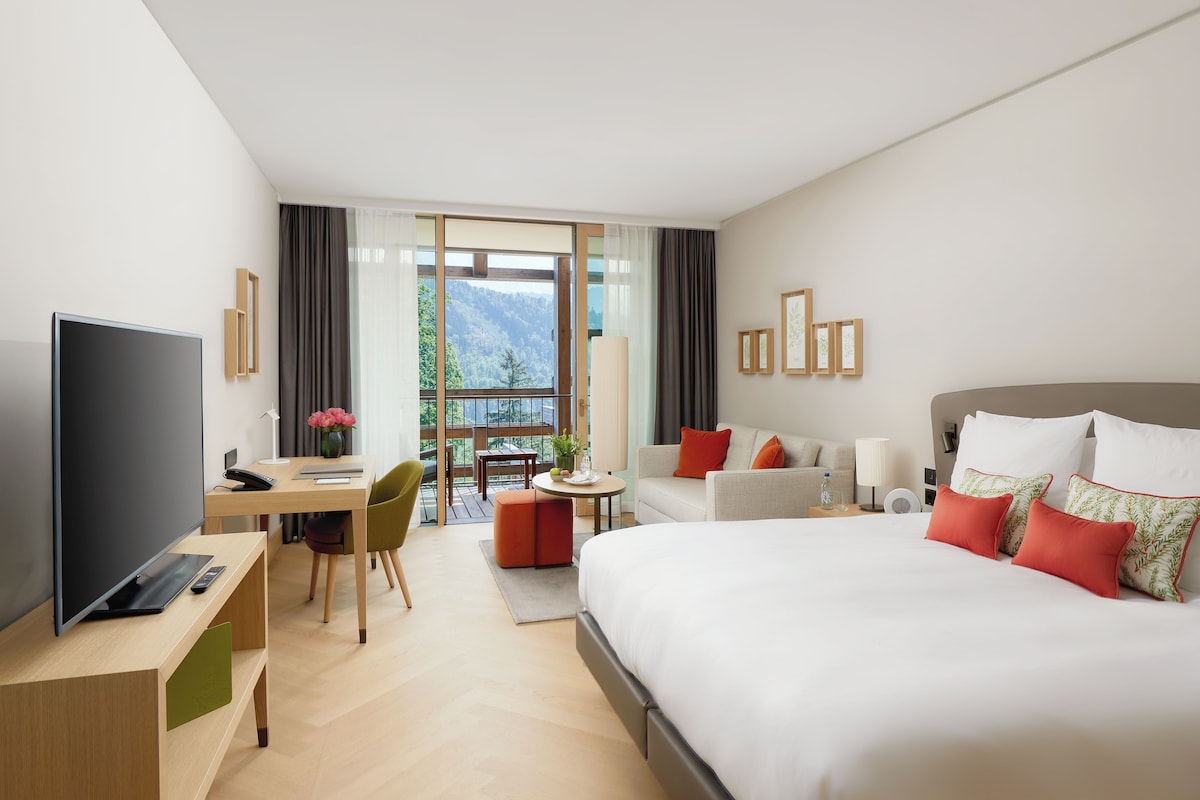 A modern hotel room with a large bed, a desk with a TV, a dining area, and a view of the outdoors.