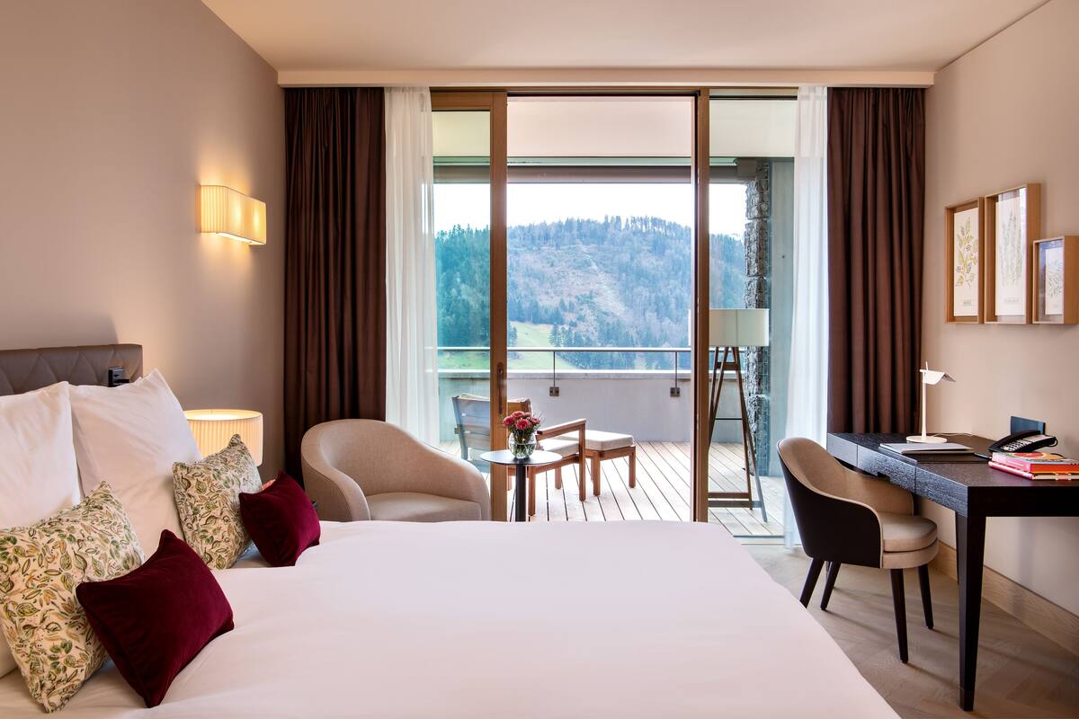 A hotel room with a large bed, a desk, a chair, and a balcony with a view of the mountains.