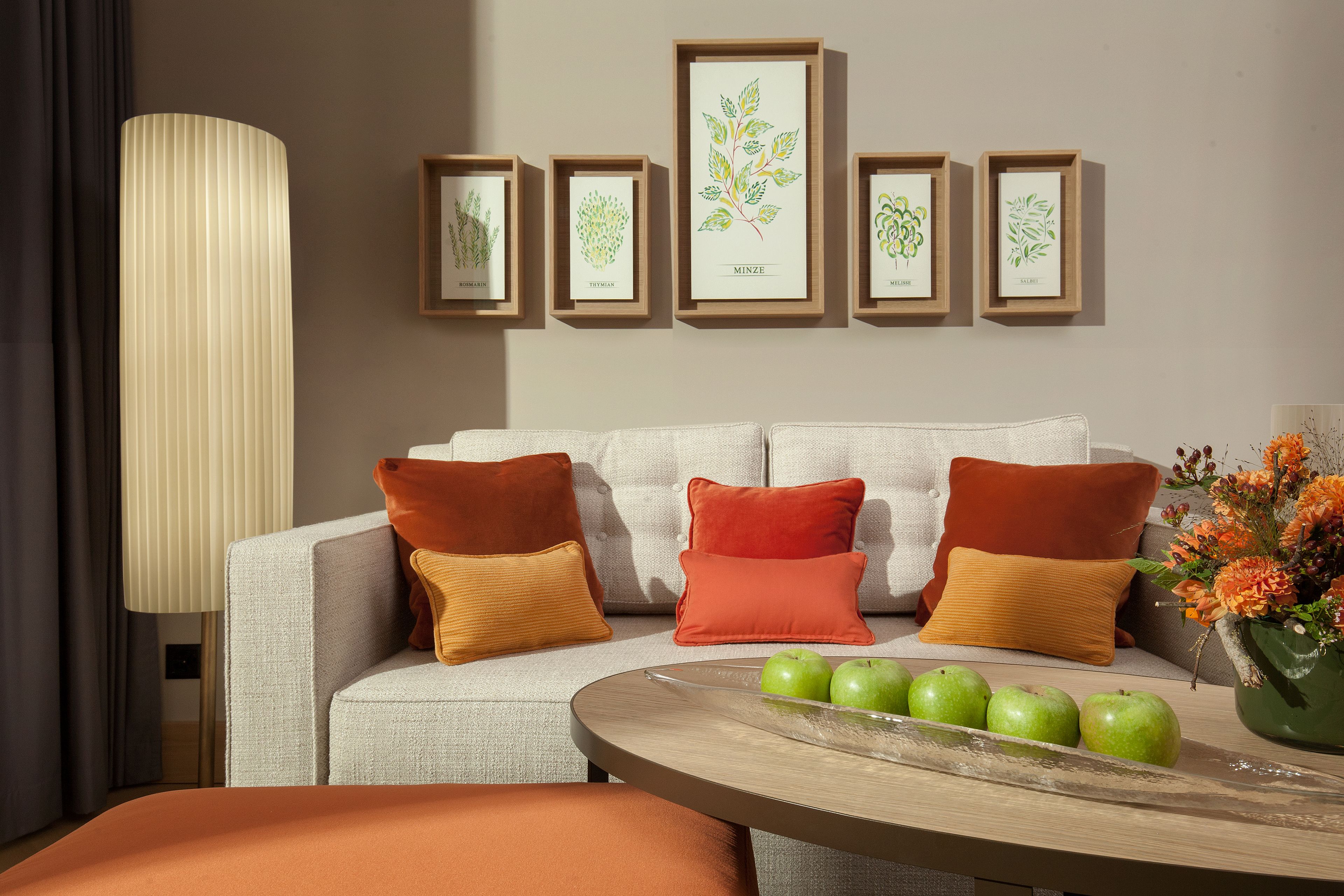 A modern living room with a sofa, pillows, framed botanical prints, a lamp, and a table with apples and flowers.