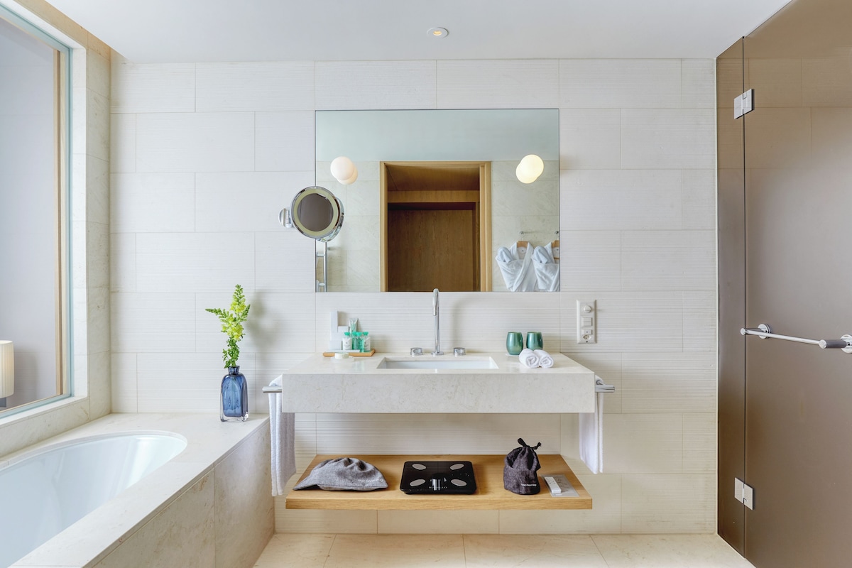 A modern bathroom with a large mirror, a sink, a shower, and a shelf with toiletries.