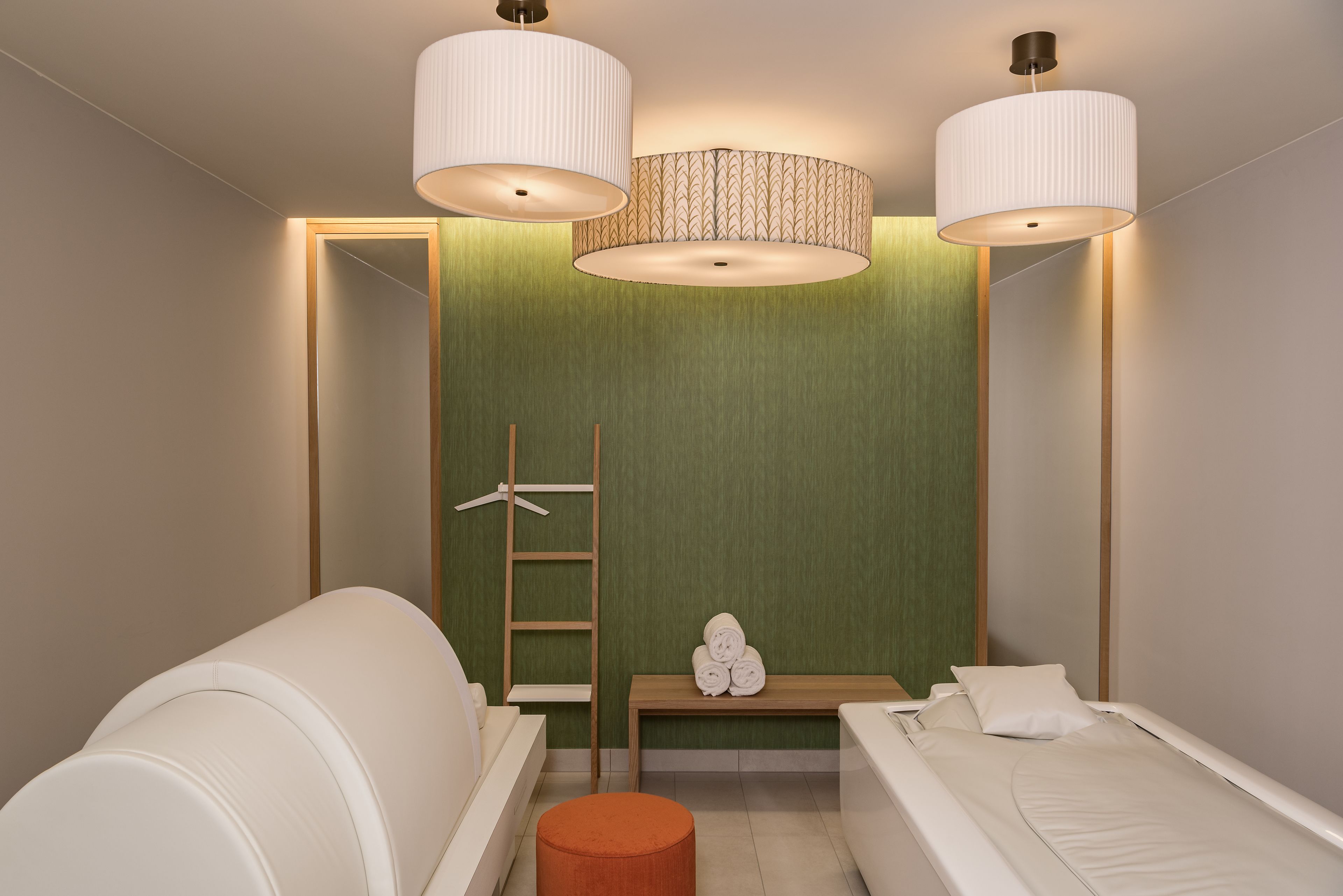 A modern bedroom with a green accent wall, two white beds, a wooden bench with towels, and a ladder.