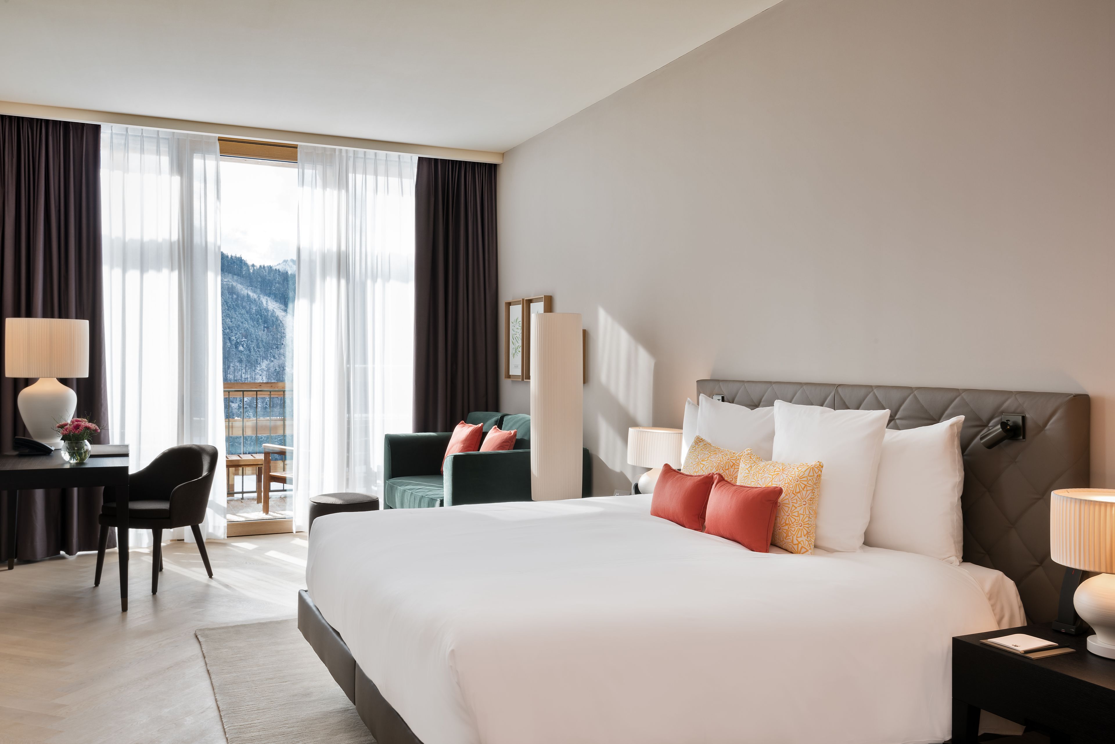 Habitación de hotel con una cama grande, un escritorio, una silla, una lámpara y vistas a las montañas a través de la ventana.