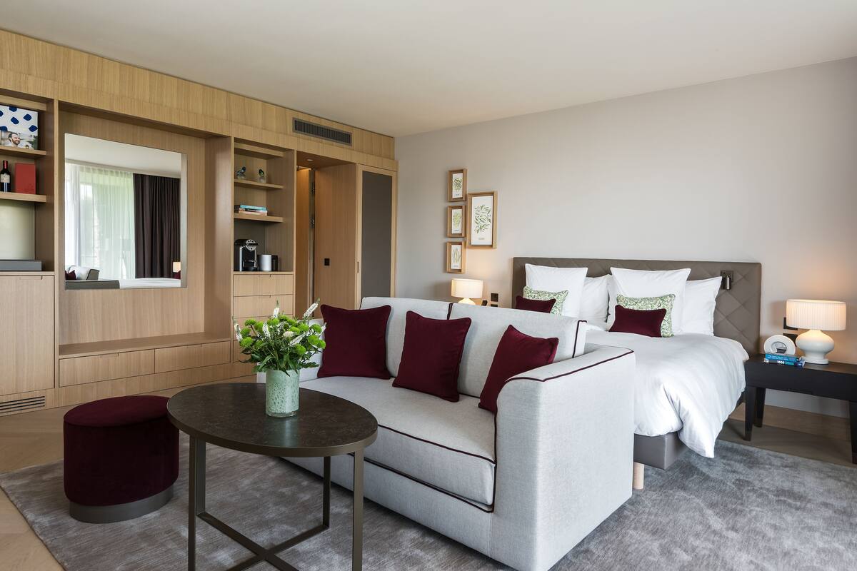 A modern hotel room with a sofa, a bed, a small table, and a bedside table with a lamp.