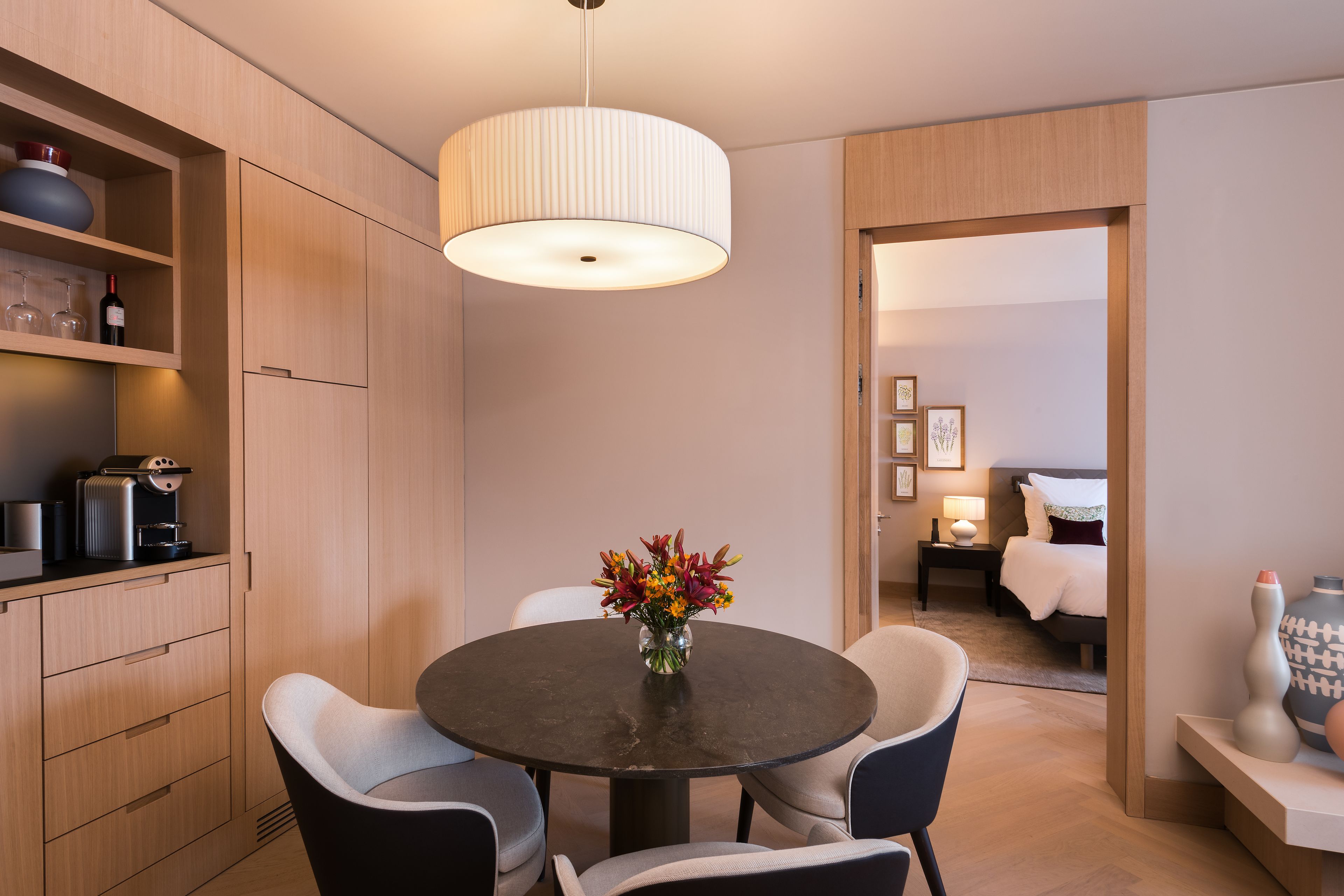 Una habitación de hotel moderna con una mesa de comedor redonda, sillas, un florero, una máquina de café y una cama con una mesita de noche.