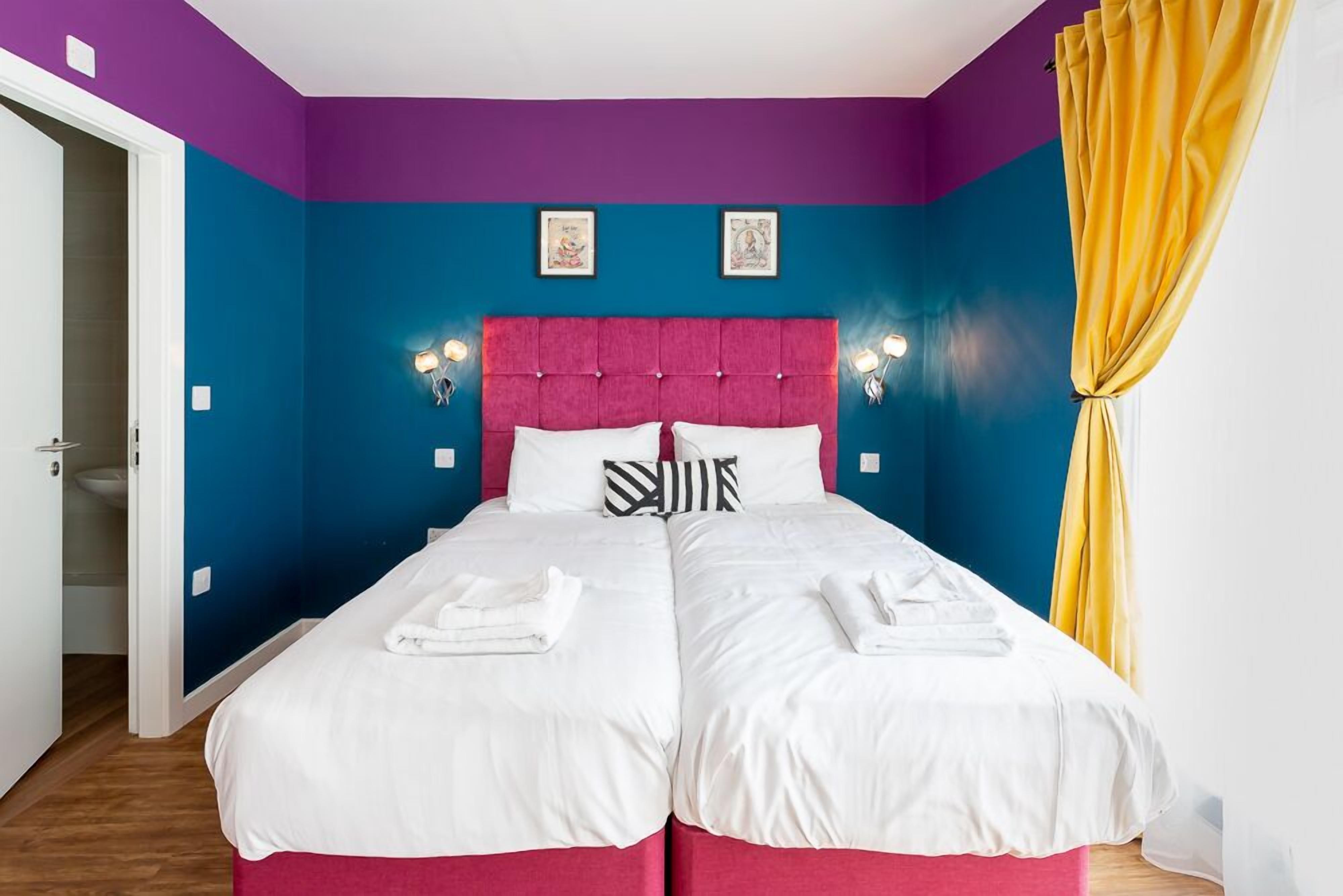 A double bed with white bedding, a pink headboard, and two wall-mounted lights.