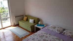 A room with a green sofa, a small white nightstand with a lamp, a floral bedspread, and a wooden floor. - Apartmani Lastavica (Orebic)