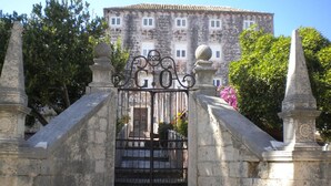 Una escalera de piedra conduce a una entrada protegida por un portón de hierro forjado, flanqueada por dos pilares de piedra con parte superior esférica.