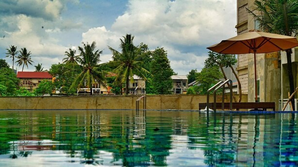 A swimming pool with clear water reflecting palm trees and buildings. - CoreHotel Malioboro City Yogyakarta (Depok)