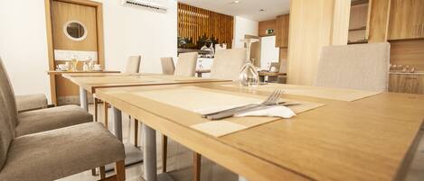 A modern dining area with a long wooden table, beige upholstered chairs, and a kitchen in the background.