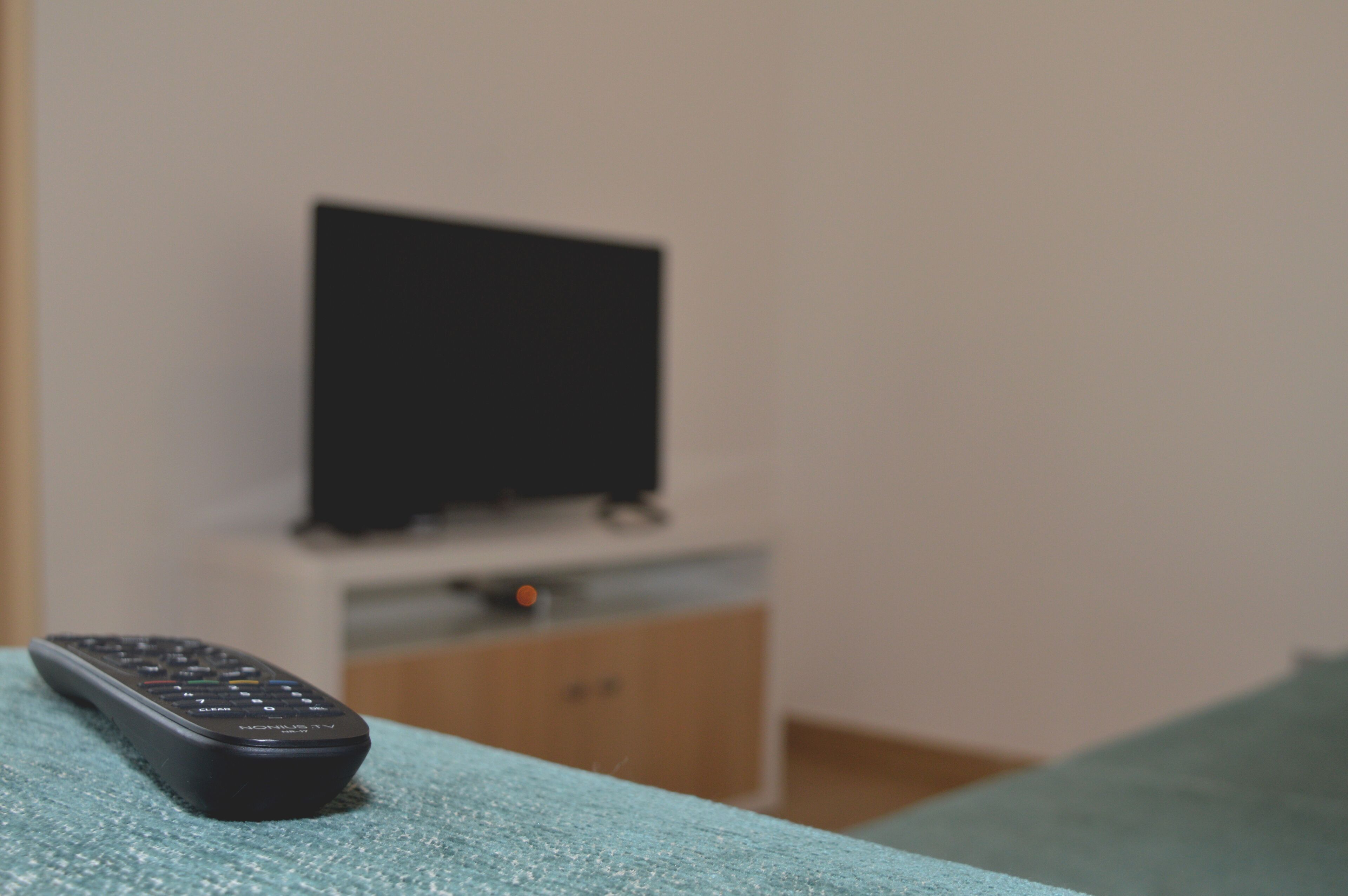 A remote control on a teal surface with a television set on a wooden cabinet in the background.