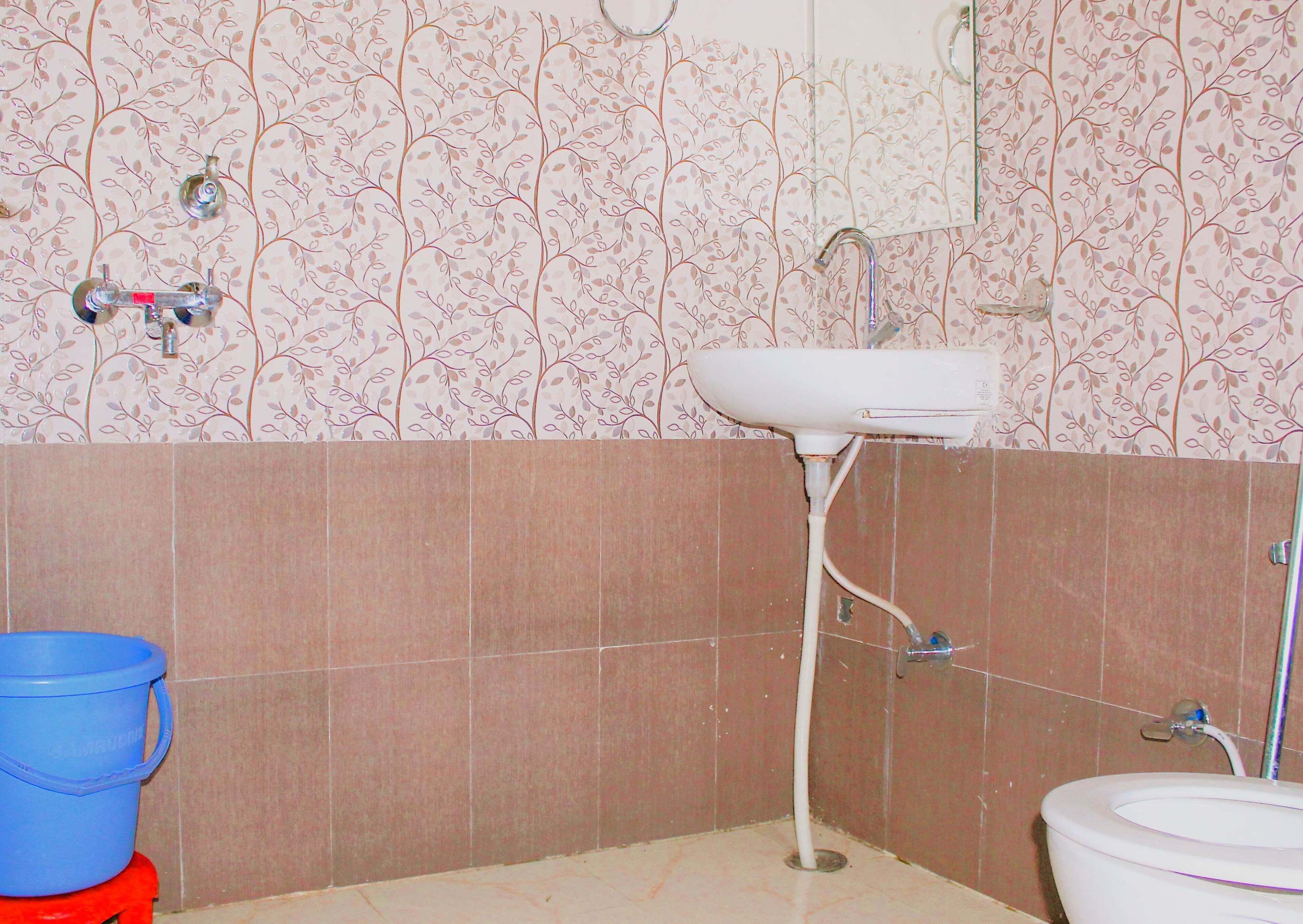 Kylpyhuoneessa on valkoinen pesuallas, peili ja wc-istuin. Seinässä on vaaleanpunaisia, kukkakuvioituja laattoja ja lattialla sininen ämpäri.