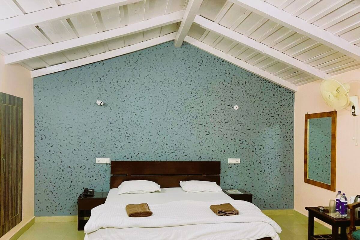 Un dormitorio con techo inclinado, una cama con ropa blanca, dos toallas dobladas, una mesita de noche y un ventilador de pared, además de un espejo.