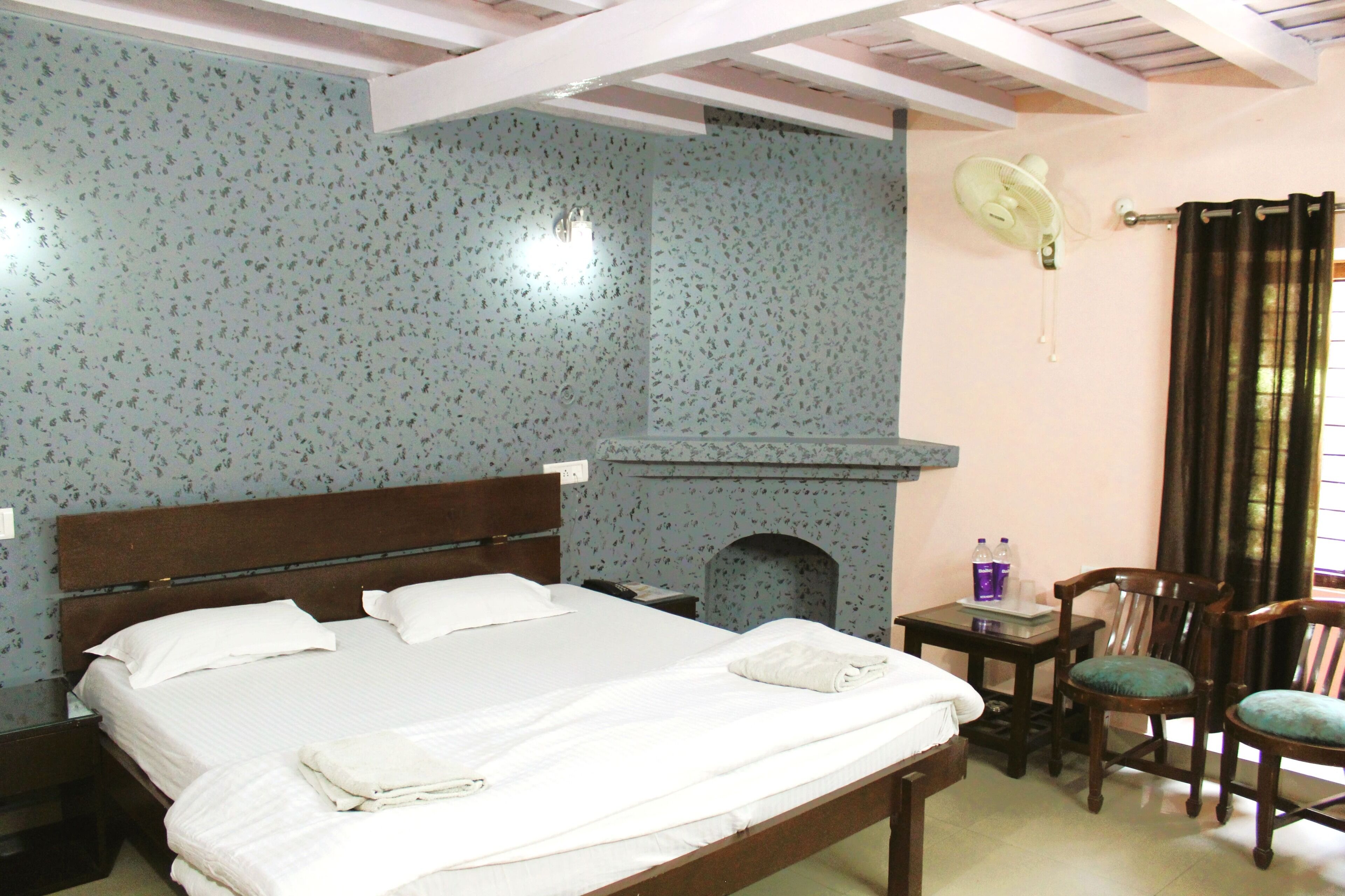 A bedroom with a bed, a small table with chairs, a wall-mounted fan, and a fireplace.