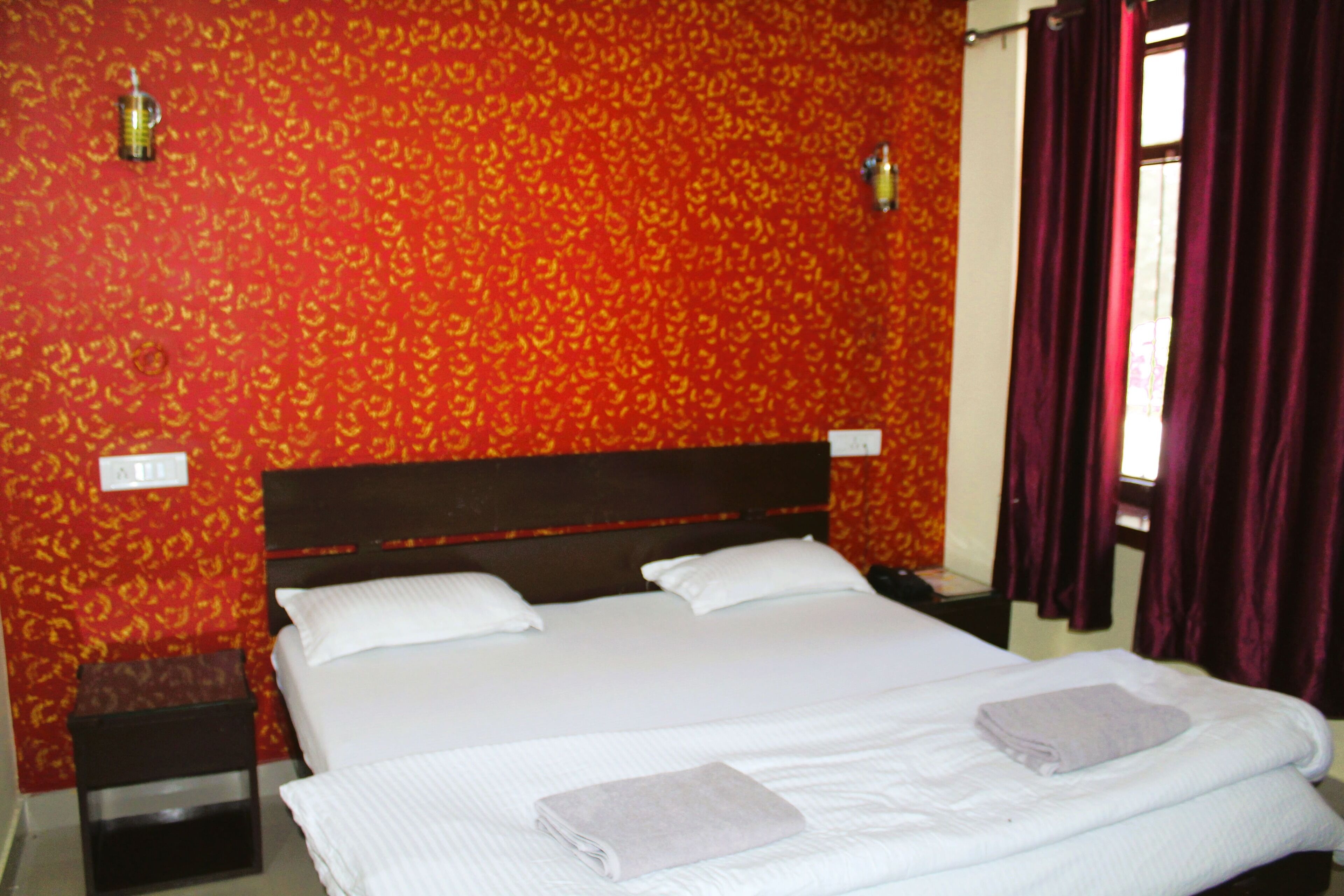 Habitación de hotel con una cama grande, dos almohadas, una mesita de noche, una pequeña mesa y una ventana con cortinas.
