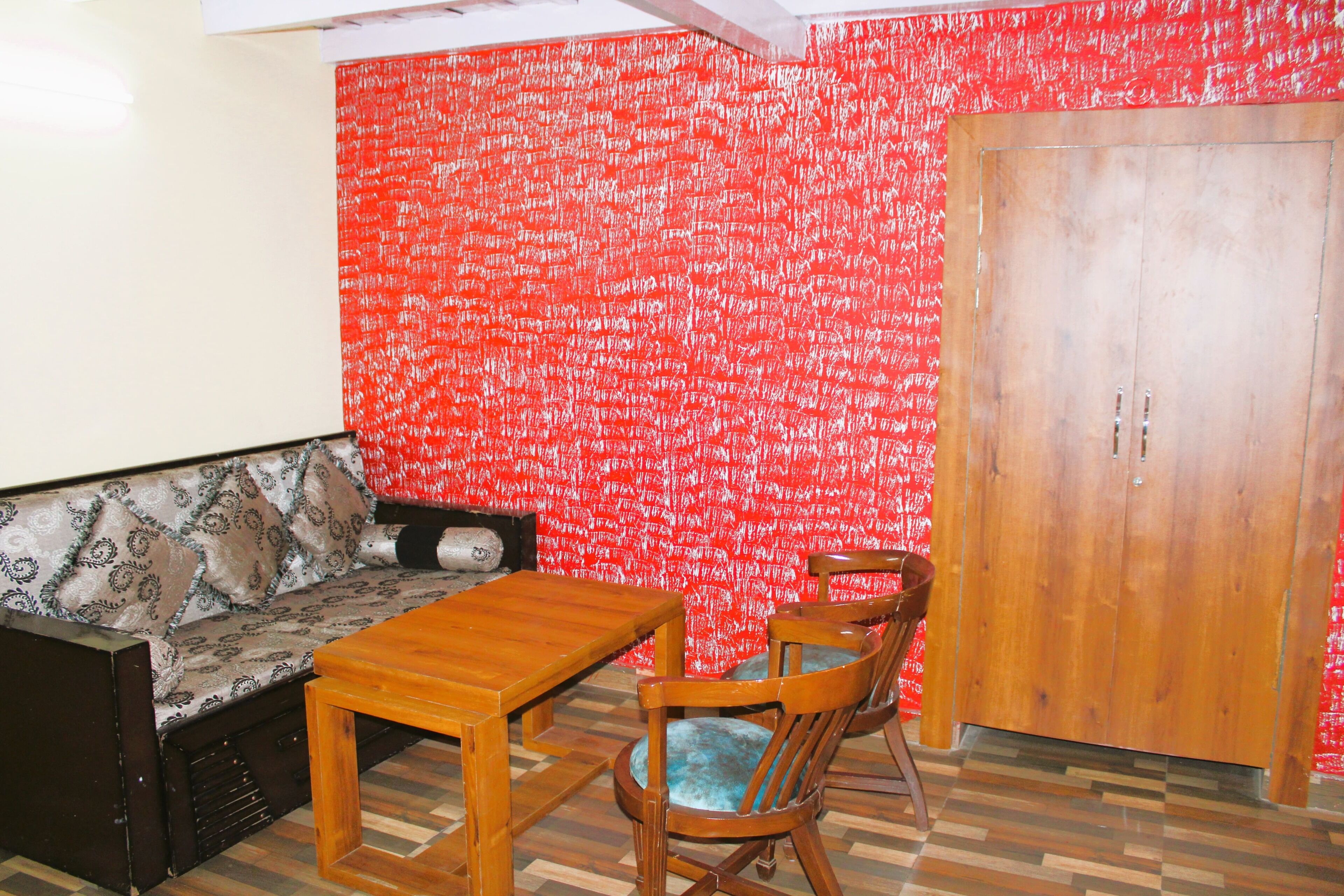 A room with a patterned sofa, wooden table, and chairs, a wardrobe, and a red patterned wall.