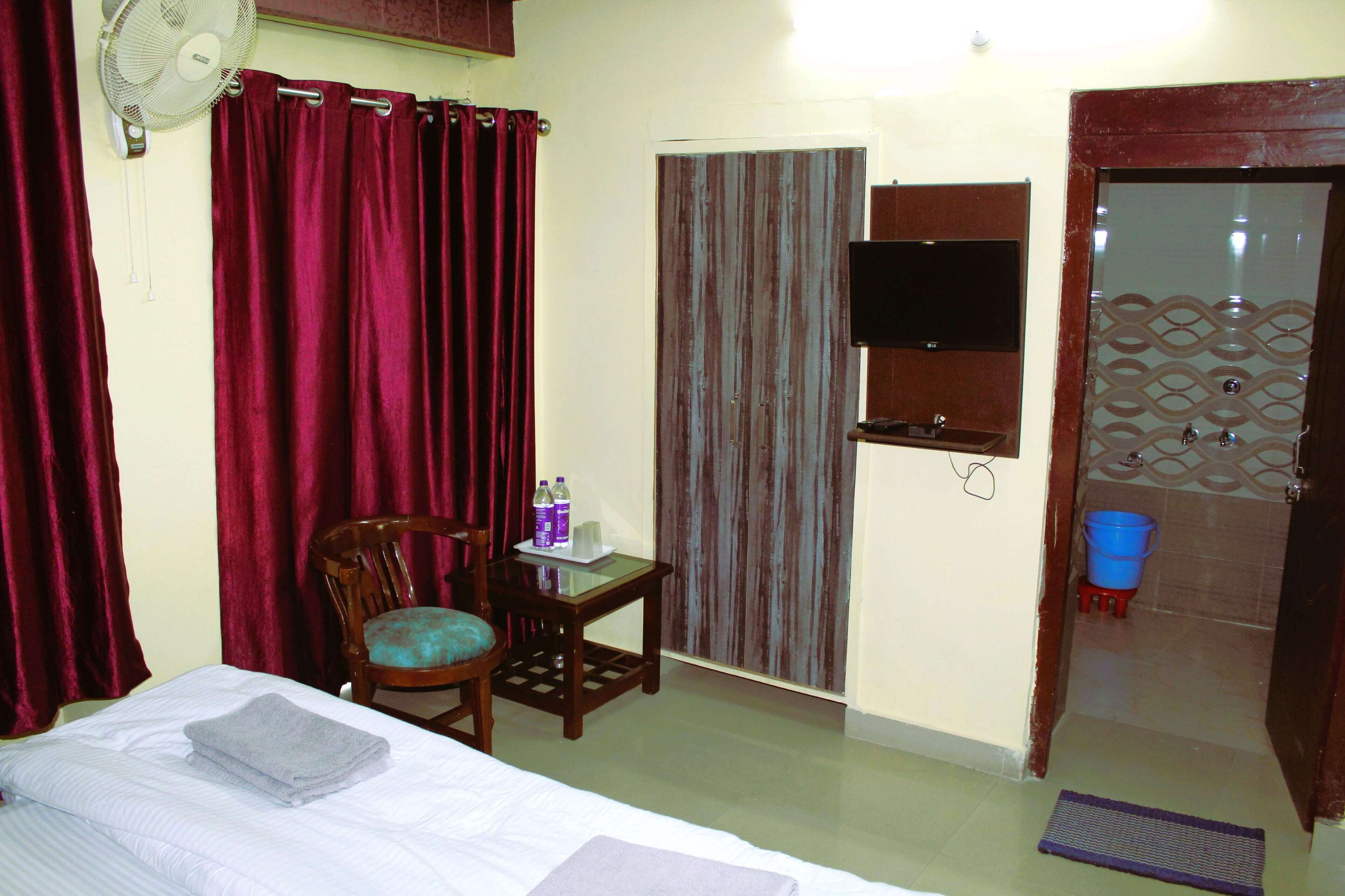 A hotel room with a bed, a chair, a small table with bottles, a fan, a TV, and a bathroom door.