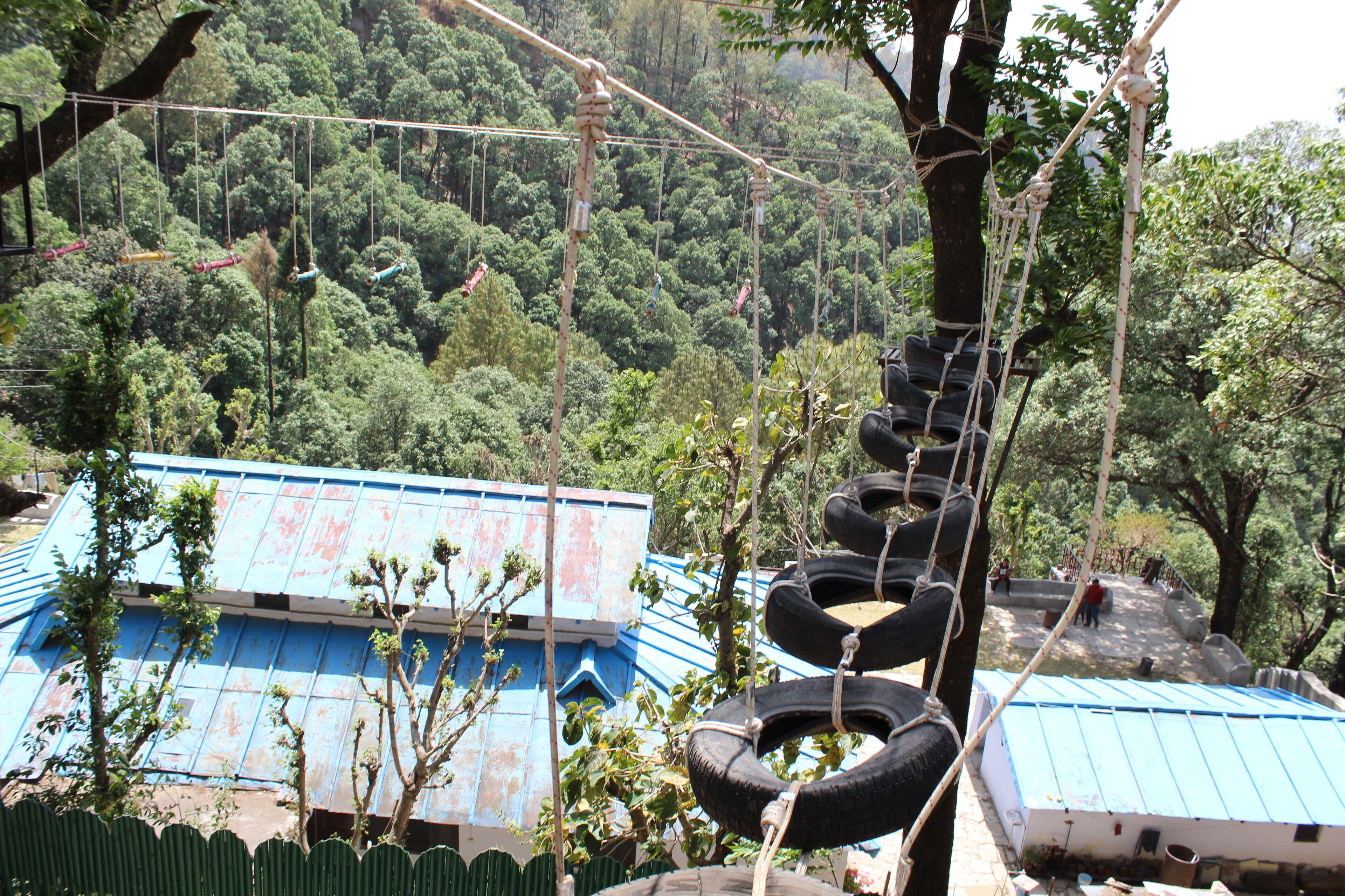A rope bridge with tires suspended above a forested area, a building with a blue roof, and a person standing on the bridge.