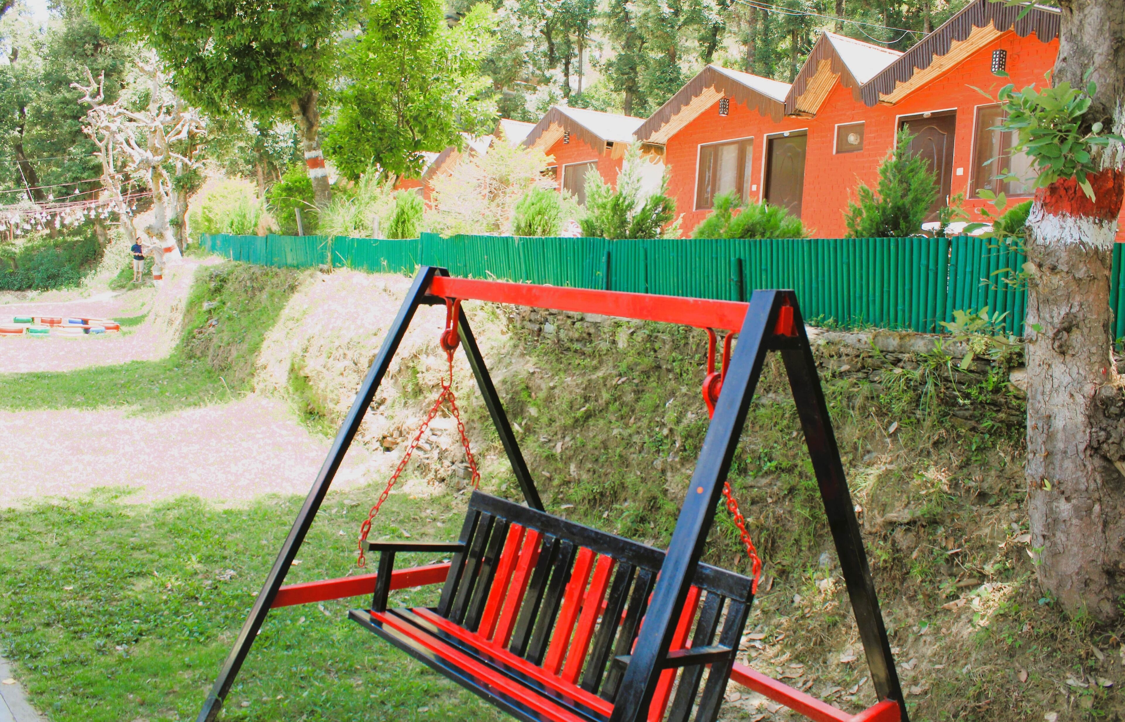 Columpios en un parque, con una cerca verde y casas rojas al fondo.